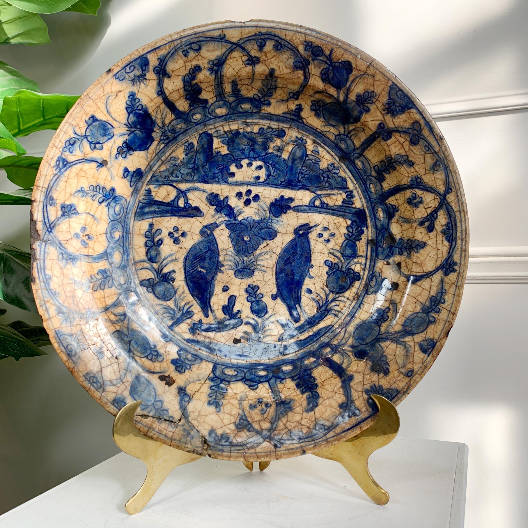 Probably Kirman, 16th/17th Century, Persia. Of Low rounded profile, on short a foot and with sloping rim, this Kirman blue and white dish with white slip decoration and stylized dense floral pattern with birds to the centre, stained and with obvious