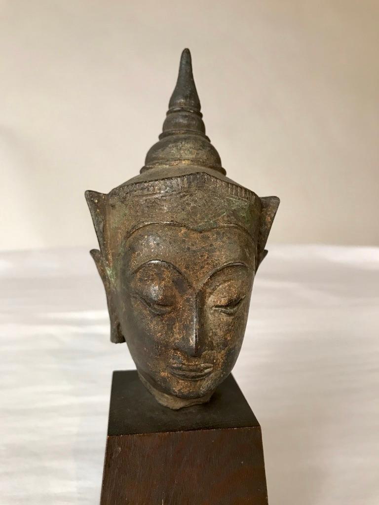 A Thai bronze head of the Buddha. Sukothai period, late 16th-early 17th century. His face with serene expression, arched eyebrows above incised eyes and lips, aquiline nose. Wearing a decorated tiara in front of a conical-shaped usnisha. Traces of