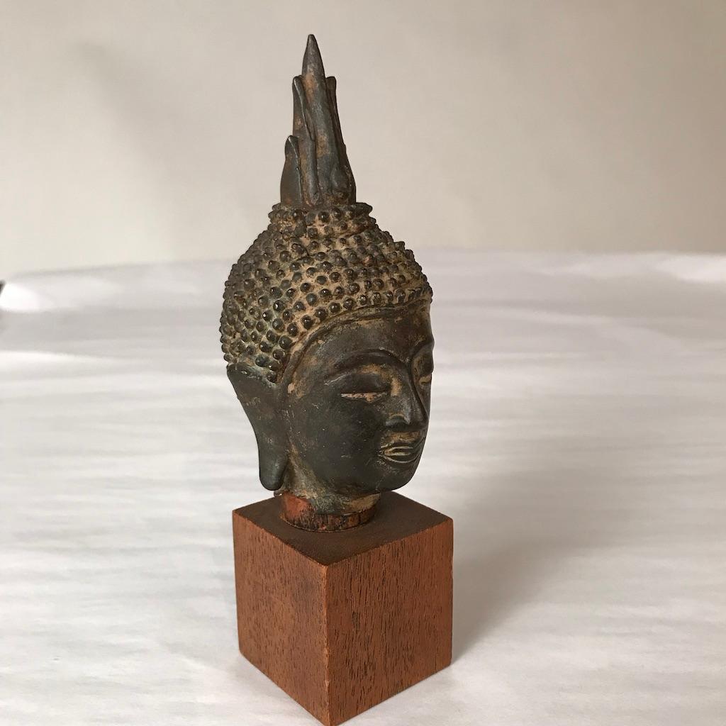 A Thai, Ayutthaya style bronze head of Buddha Shakyamuni, 16th-17th century. His face displaying serene expression, downcast eyes, below arched eyebrows, smiling lips, with curled headdress and ushnisha surmounted by a flame. Mounted on a square