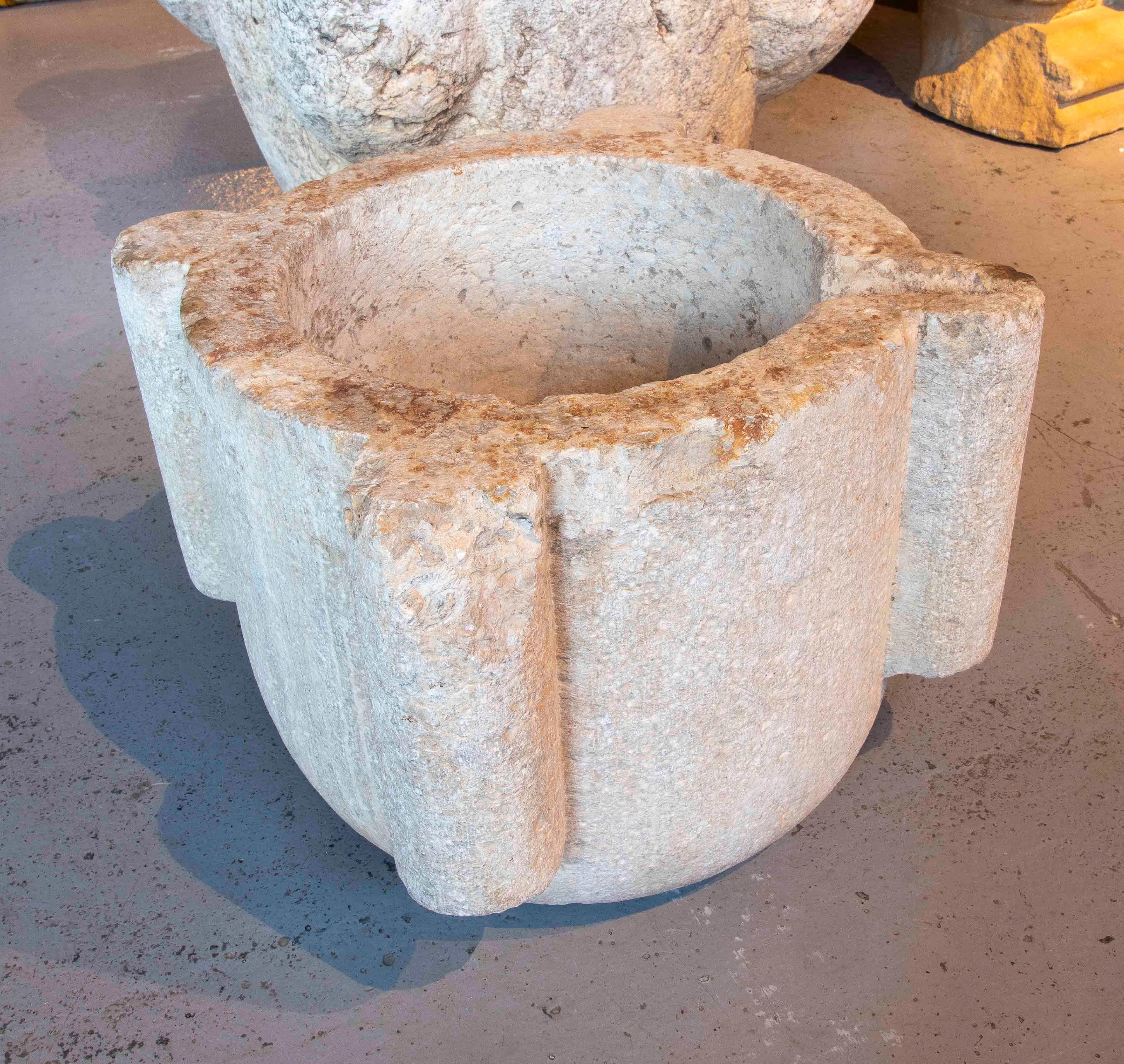 An impressive antique mortar, hand-carved from a single block of solid stone, with a cylindrical shape and sturdy vertical buttresses that reinforce its structure. Its deep cavity reveals its original use for grinding grains, herbs, and spices, both