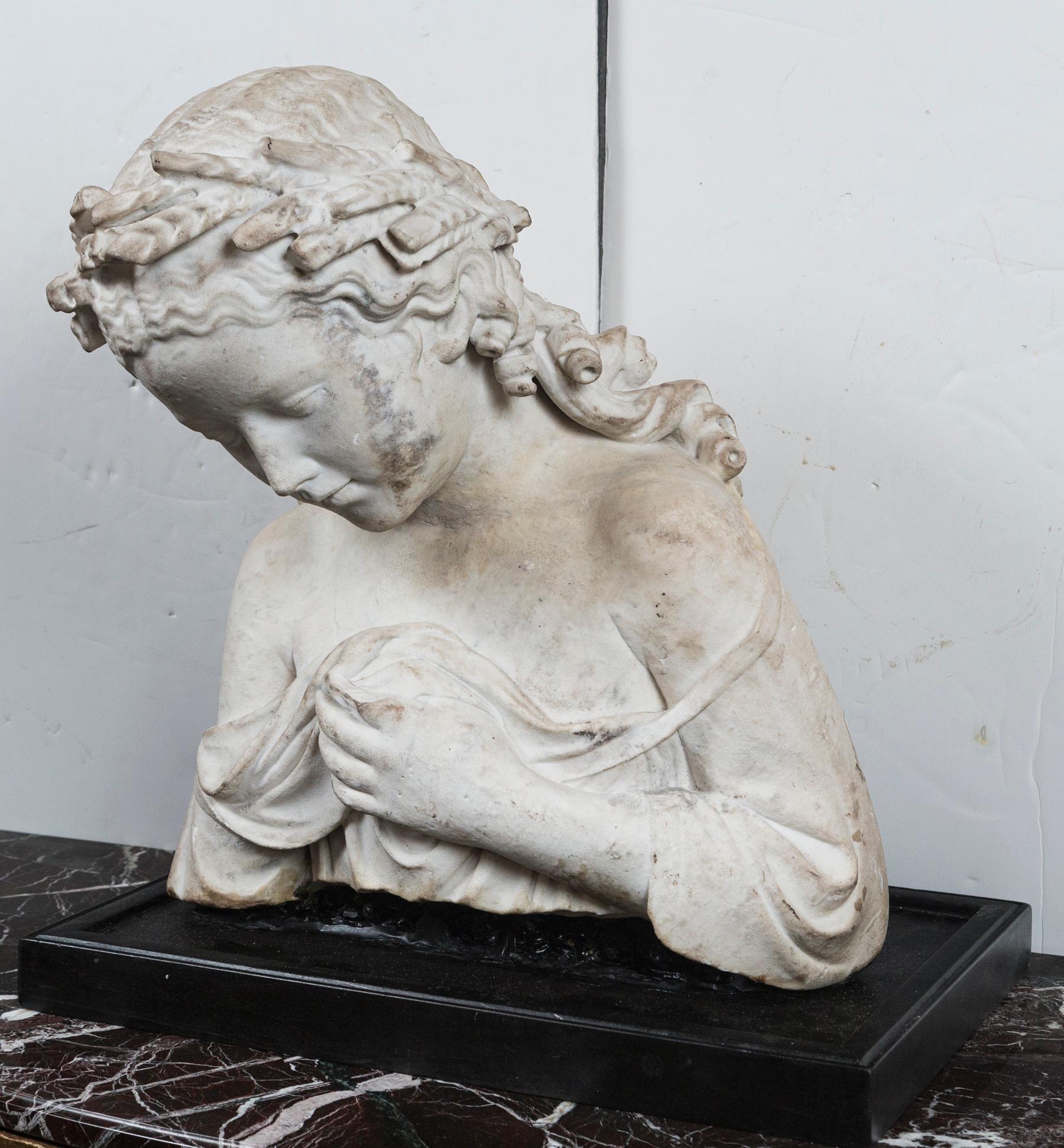 This bust is a fragment of a full figure marble statue of a woman. A modern base has been made for it.
She wears classical garb. Her left hand seems to be holding up the garment over her breast. Her right arm is missing. Her head bent forward which