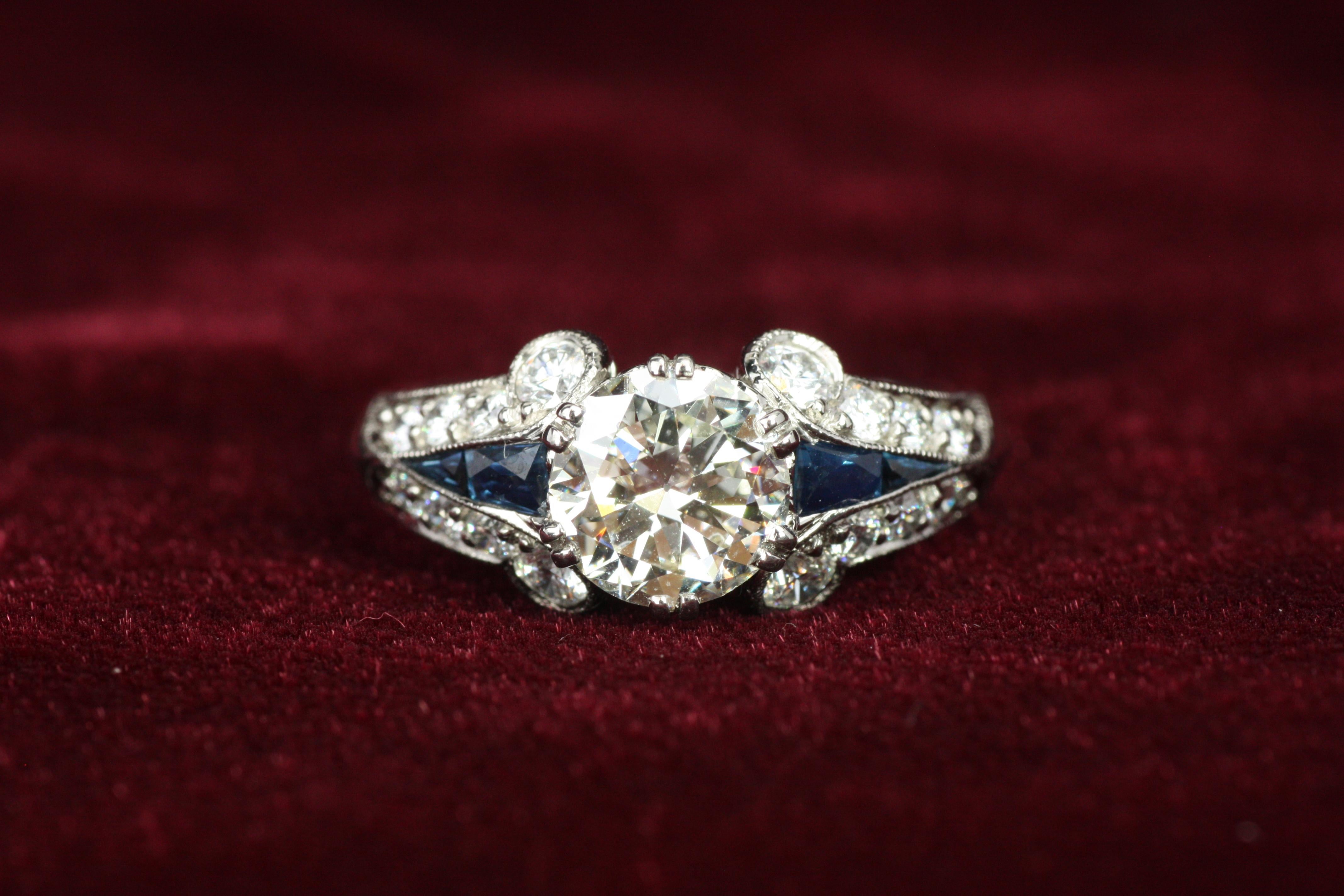 This stunning Art Deco-inspired ring is a masterpiece of vintage charm and luxurious details. Featuring a 1.14ct transitional cut center diamond, it is flanked by 16 additional sparkling diamonds and four vivid blue sapphires. Crafted in palladium,