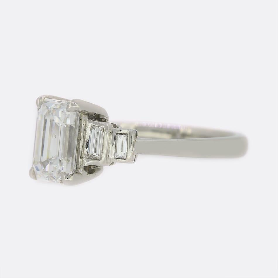 This is a platinum diamond Art Deco style engagement ring. The focal point of this ring is a certified 1.70 carat emerald cut diamond which sits claw set at the centre of the piece with a pair of additional emerald cut diamonds sitting on either