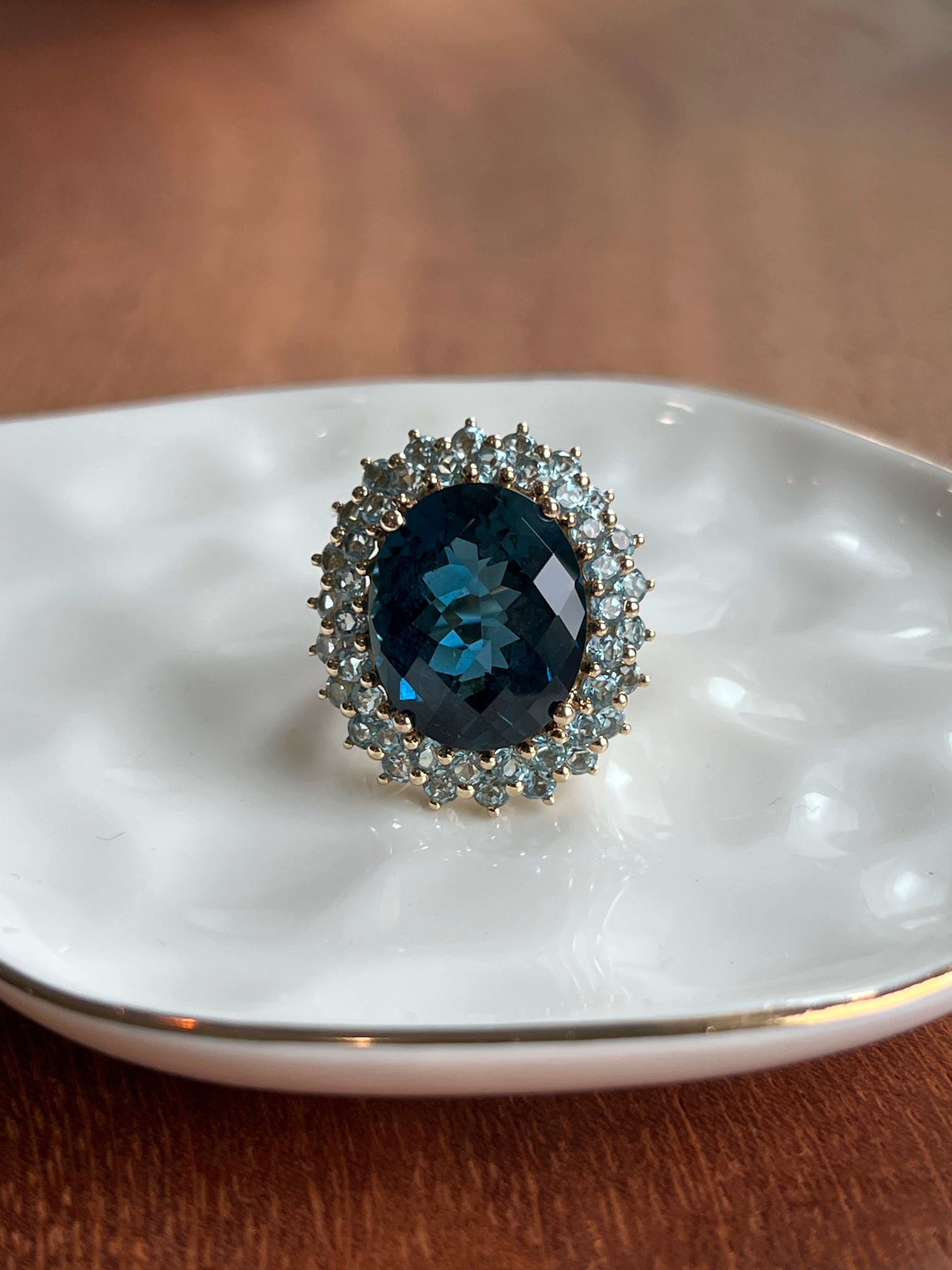 Striking and sculptural, this bold 14 karat yellow gold contemporary cocktail ring centers on an impressive 17.07 carat oval checkerboard-cut London blue topaz, prized for its deep, moody hue and mesmerizing light play. Surrounding the center stone