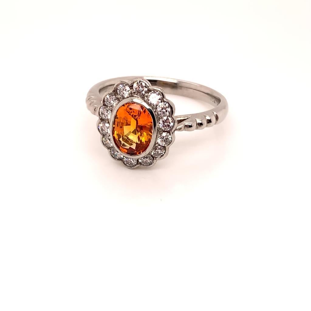At the centre of this Stunning ring is a Lustrous Oval Brilliant Orange Sapphire weighing approximately 1.72 carats. Set in 18k white gold and surrounded by approximately 0.34 carats of glittering Round Brilliant Diamonds, this eye-catching ring is