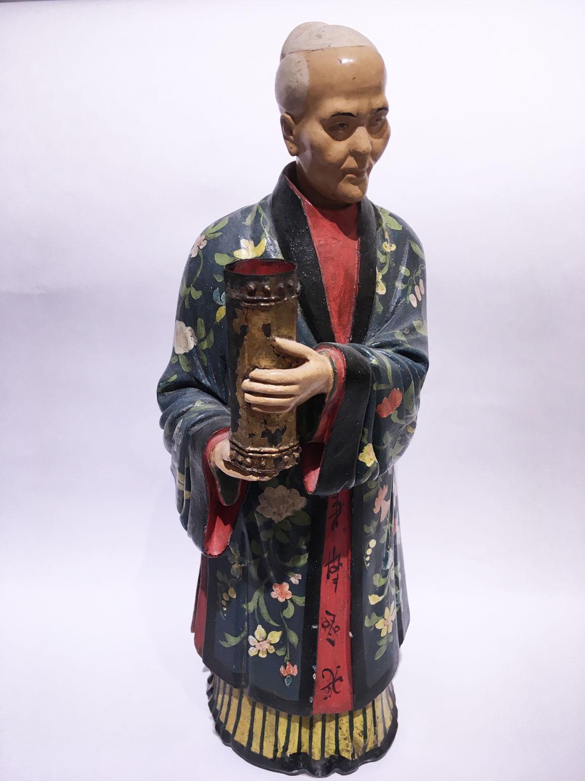 Se trata de una rara figura china de terracota lacada en  policromada con decoración floral sobre fondo verde.  Tiene el cabezal basculante. En sus manos hay un pequeño jarrón para contener plumas o plumillas de escritura. Esta sorprendente pieza