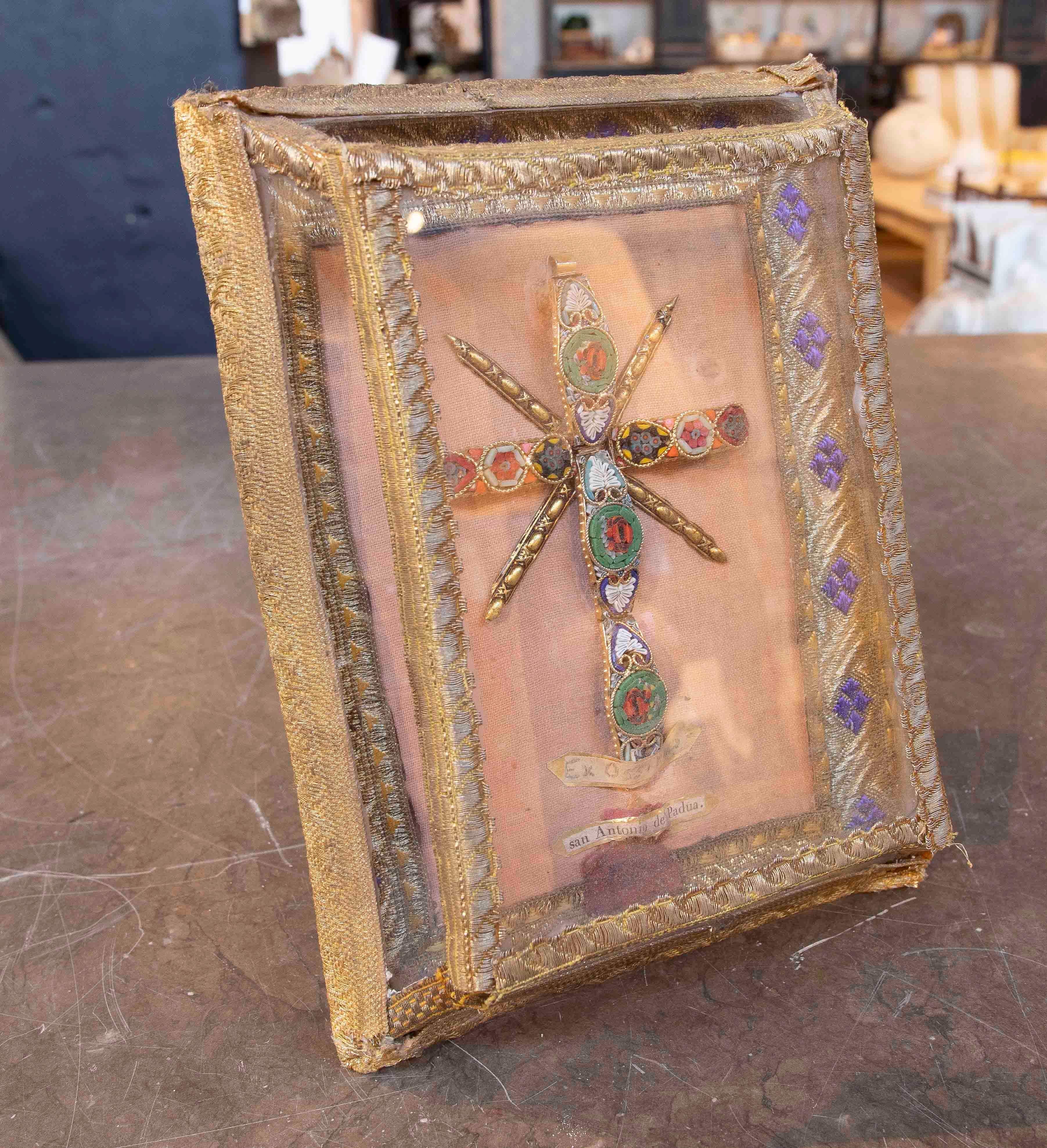 Exquise relique religieuse de Saint Antoine de Padua, protégée dans un délicat cadre en tissu brodé avec des détails dorés. Au centre, une croix décorée de micromosaïques dans des tons verts, rouges et bleus rehausse sa valeur artisanale et