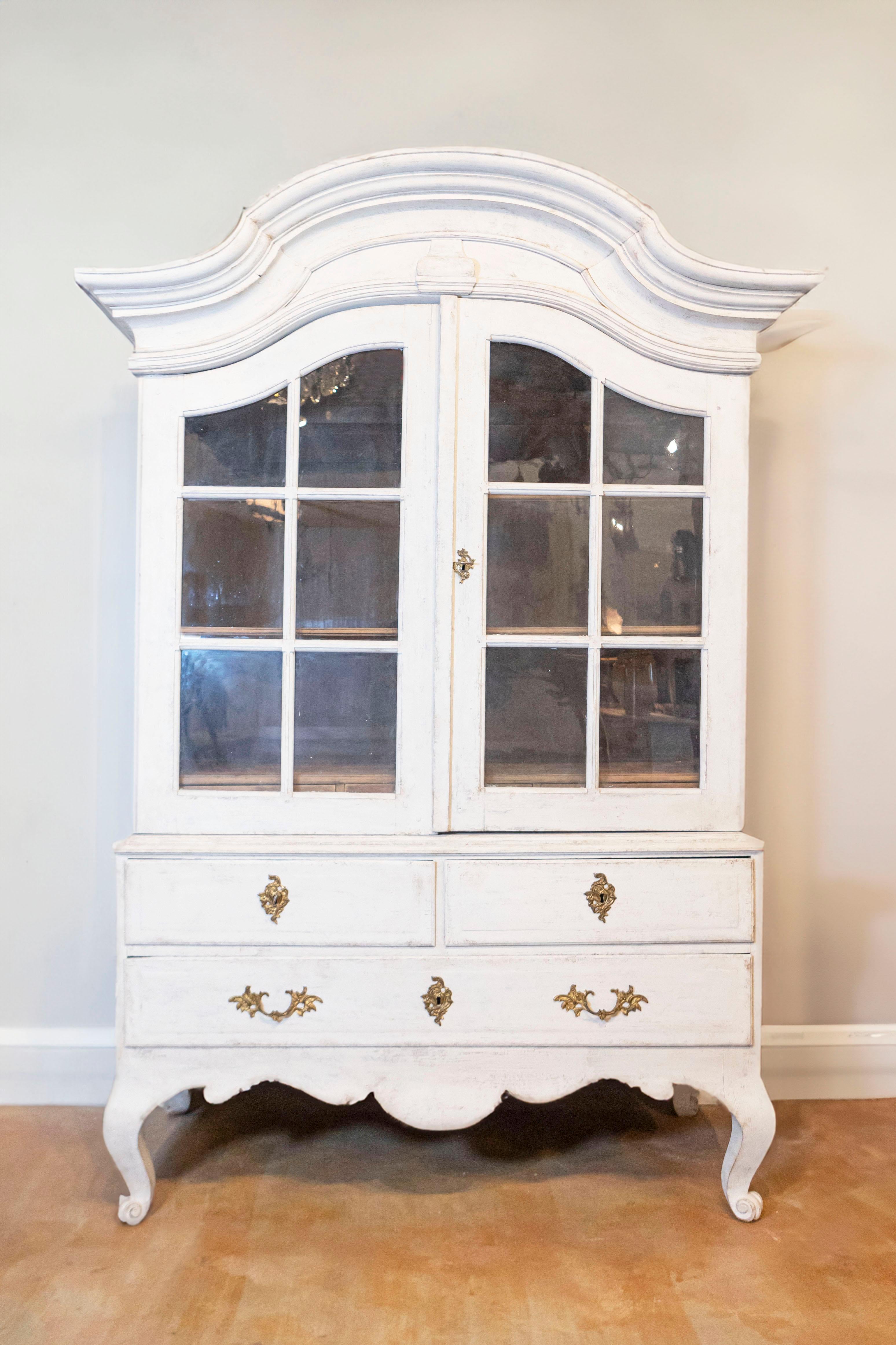 A Swedish period Rococo painted wood cabinet with bonnet-shaped pediment, glass doors, lower drawers and original hardware from the mid 18th century. Our eye is immediately attracted to the curvy molded bonnet top, beautifully crowing the confirming