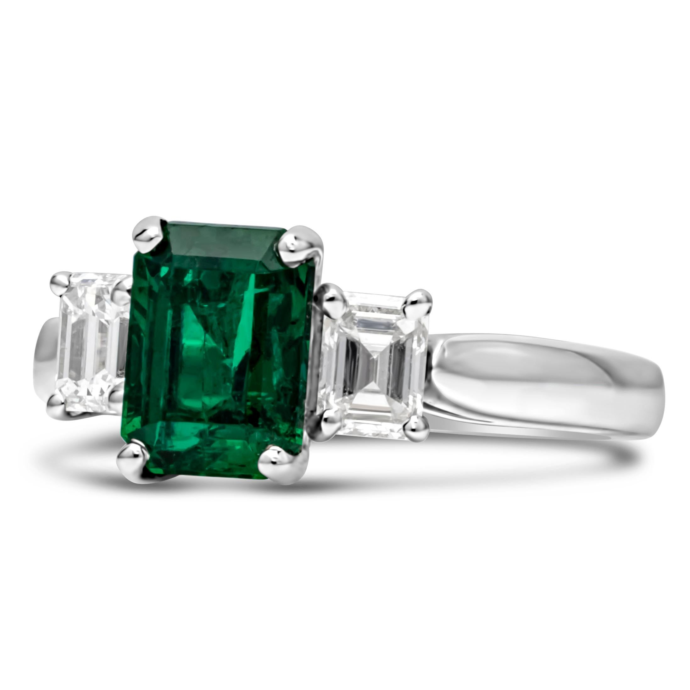 A color-rich three stone engagement ring showcasing a 1.78 carats emerald cut green emerald, set in a classic four prong basket setting. Flanked by two emerald cut brilliant diamond on each side weighing 0.70 carats total, set in a four prong basket