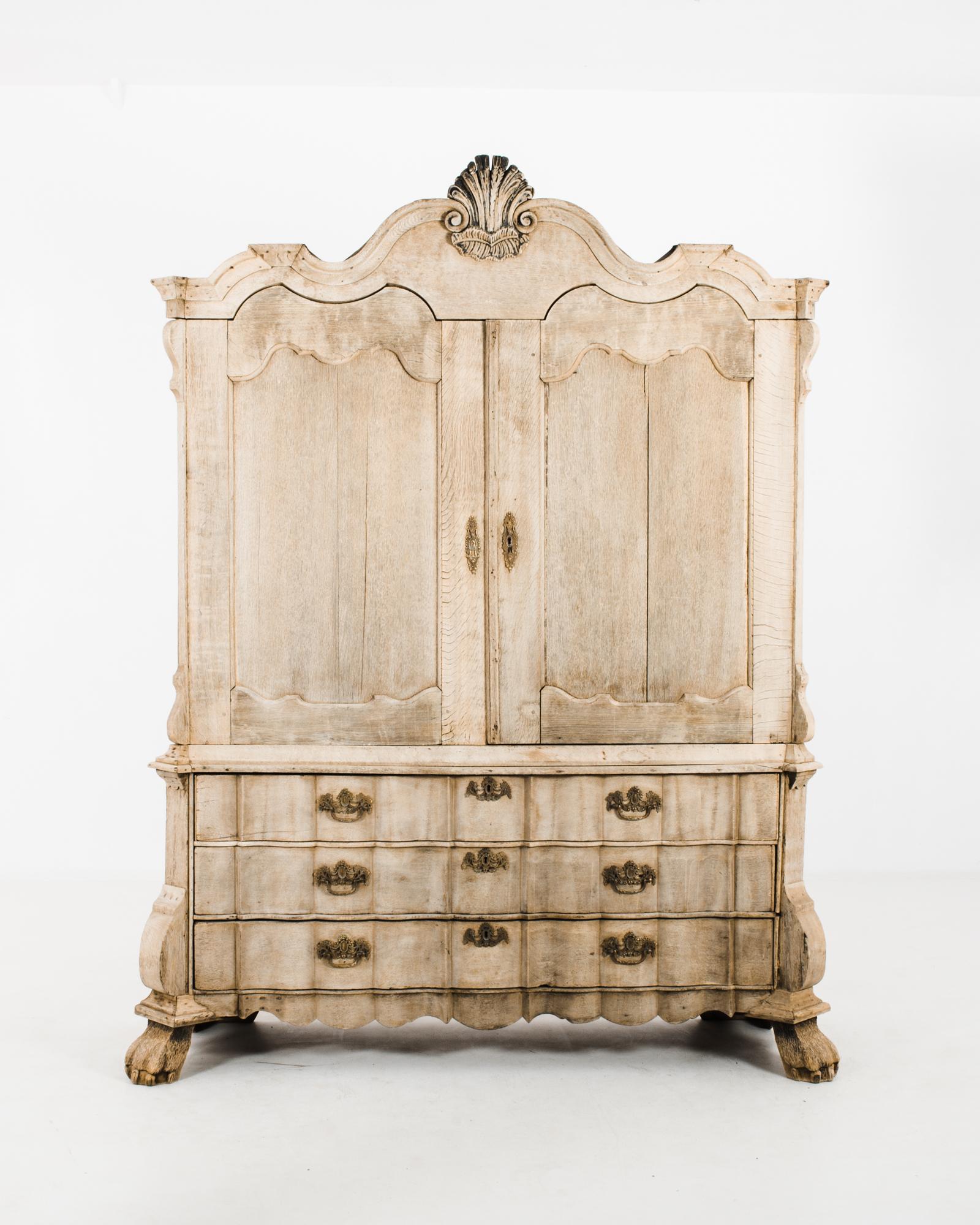 Cette armoire en chêne blanchi a été fabriquée aux Pays-Bas, vers 1780. Il présente un écusson orné, très répandu dans le mobilier hollandais des XVIIIe et XIXe siècles, avec une coquille Saint-Jacques et des feuilles. Le meuble supérieur se compose