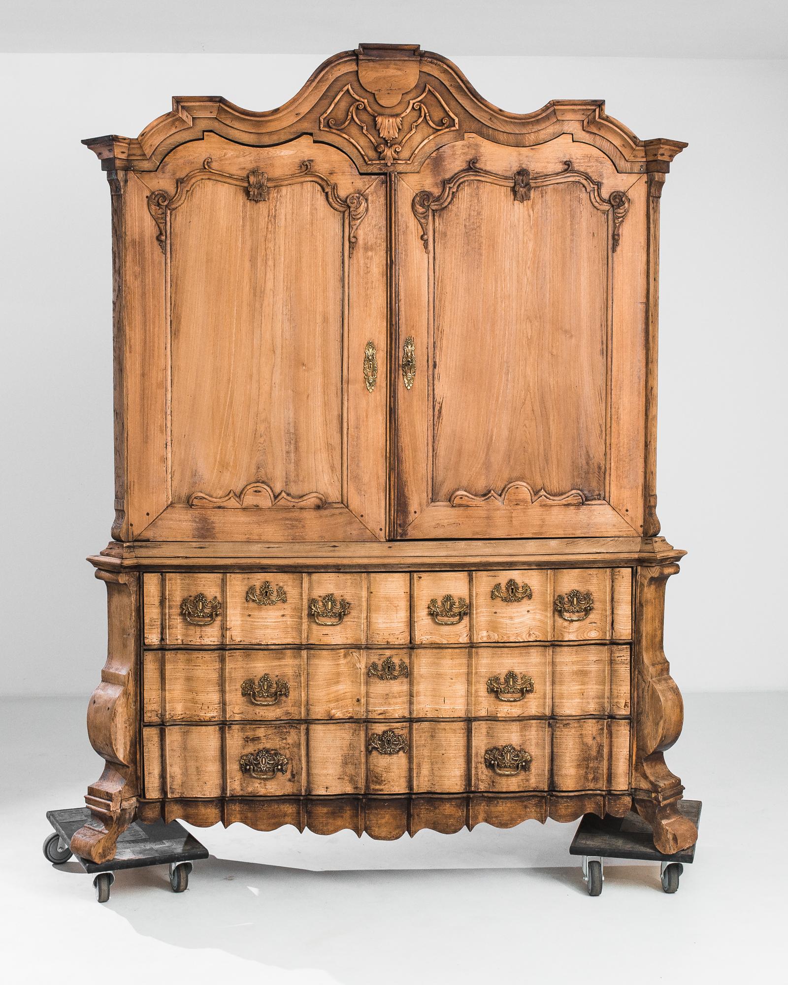 This 1780s Dutch Wooden Cabinet is an exquisite specimen of the refined craftsmanship from the period. Its robust wooden structure is adorned with ornate carvings and a curved bonnet top that exudes the grandeur of the era. The warm, natural patina
