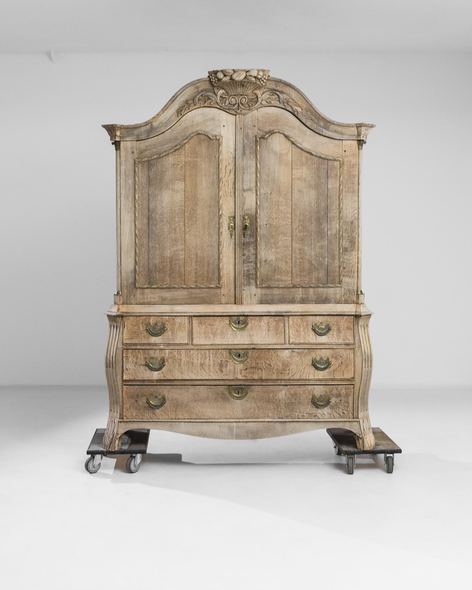 An 1780s Dutch bleached oak cabinet. This grand cabinet offers three sliding bottom drawers and a large top compartment fit with subsequent, smaller drawer pulls. A scrupulously hand-carved detail of a cornucopia sits atop it, while roman motifs