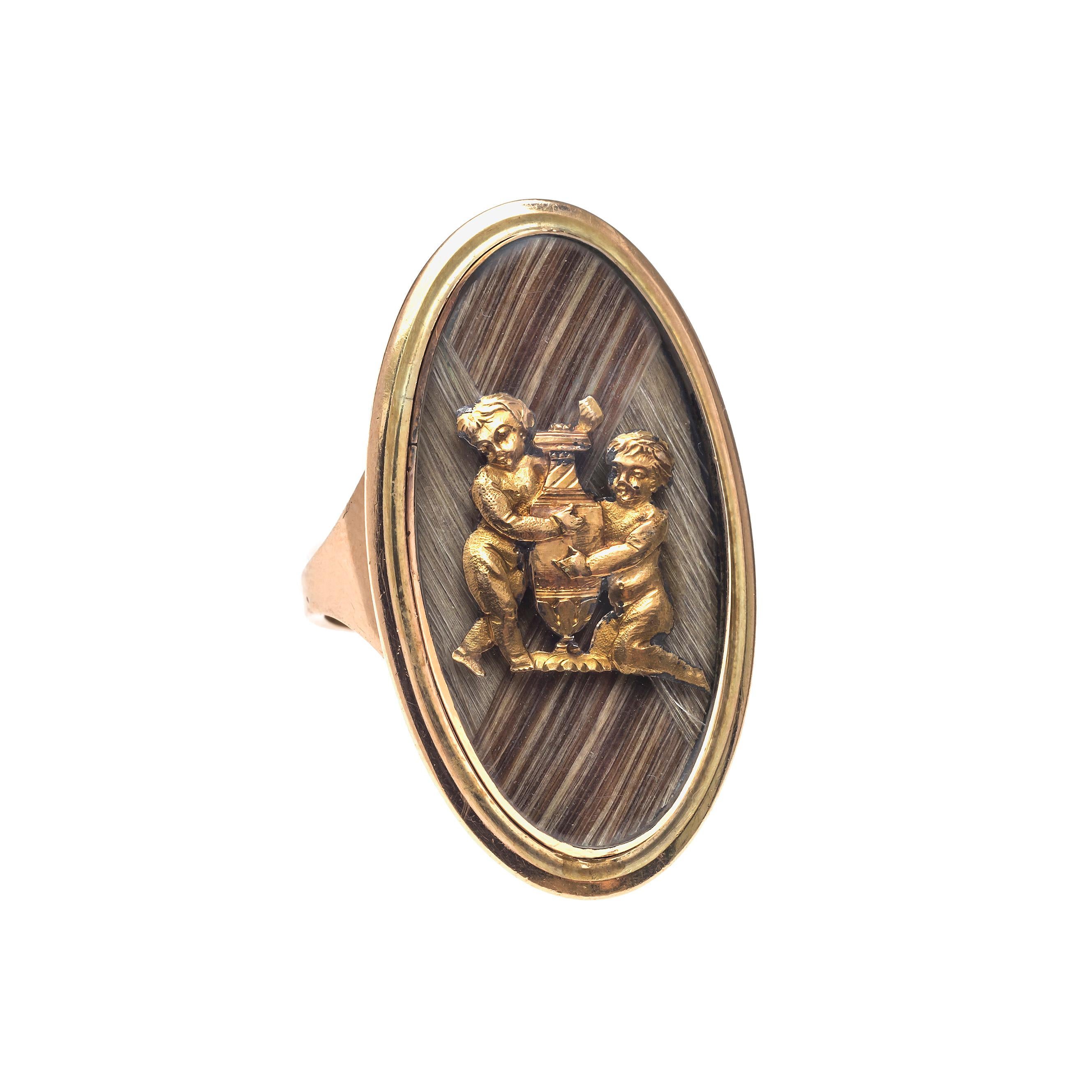 The oval 14K gold ring is typical of the style of the last decade of the 18th century. Two different kinds of hair have been interwoven and decorated with two finely engraved golden putti that are embracing an amphora. The ring is most likely the