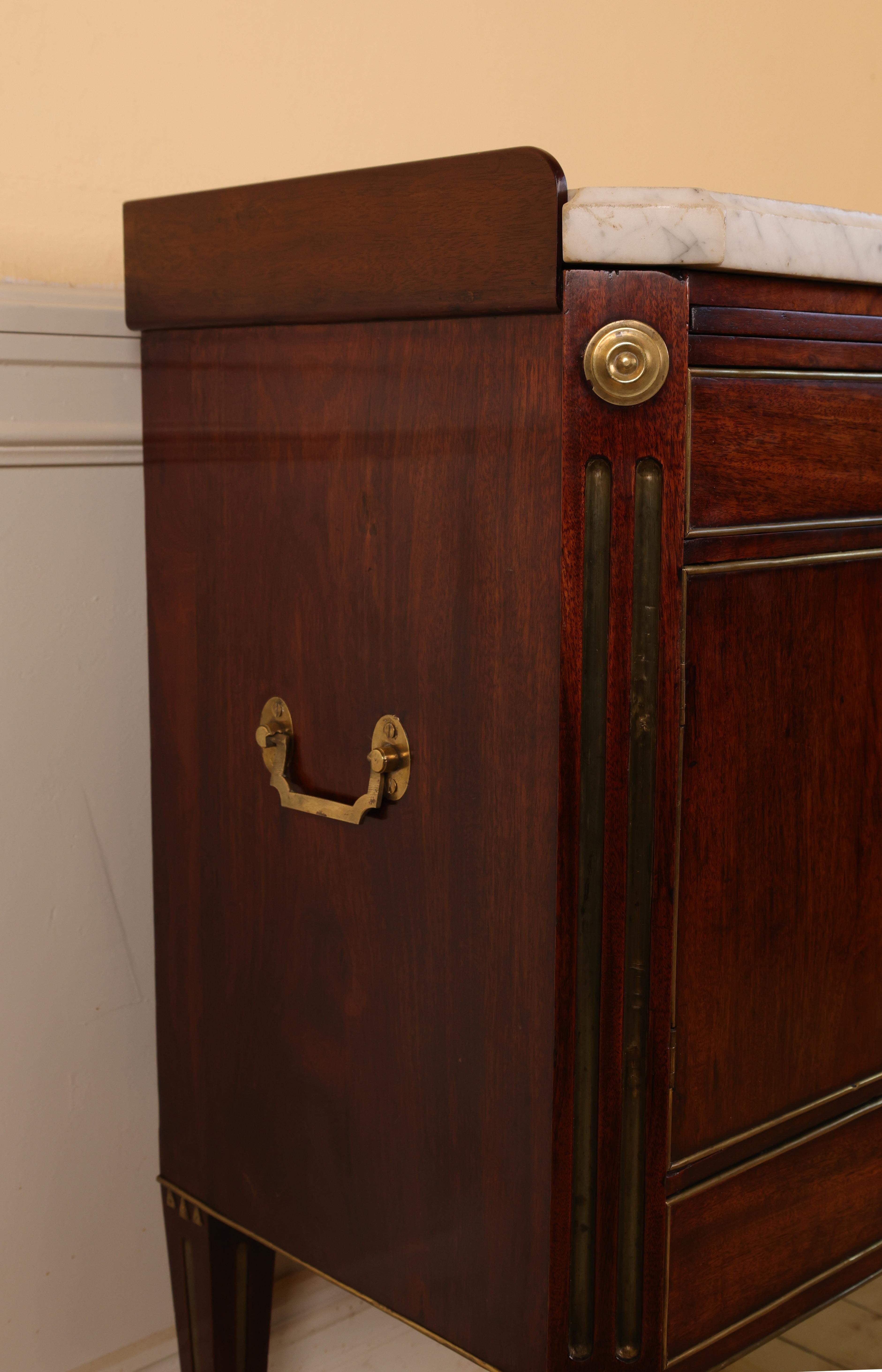 A late-Gustavian nightstand made in Stockholm, circa 1790. The cabinet features angled side corners with fluting and is veneered in mahogany. Rich brass moldings frame the doors and drawers. The original Carrara marble top remains intact. The sides
