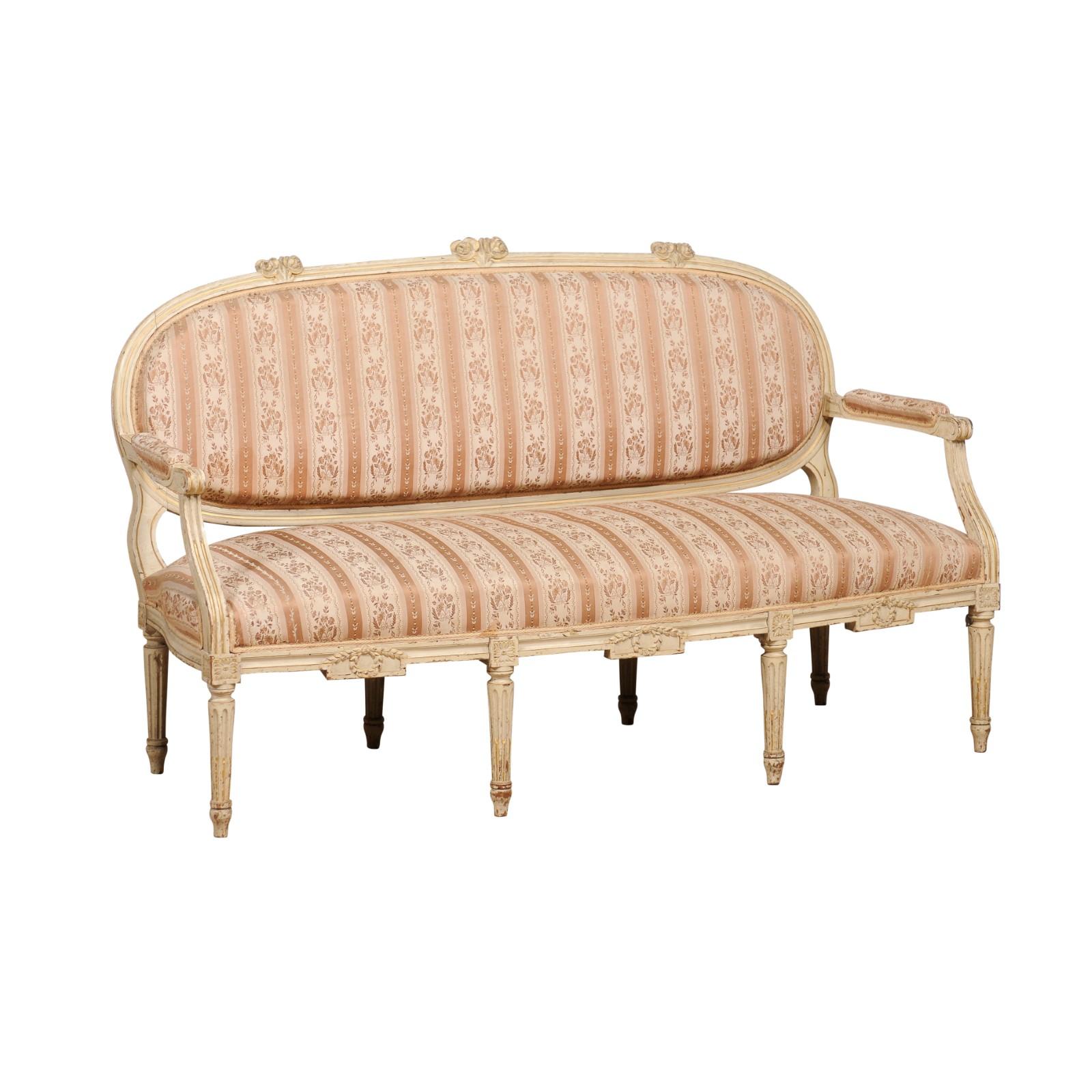 A French Louis XVI period sofa from circa 1790 with antique beige painted finish, oval back, carved foliage and fluted legs. Indulge in the romance of bygone eras with this exquisite French Louis XVI period sofa, circa 1790. Showcasing an antique