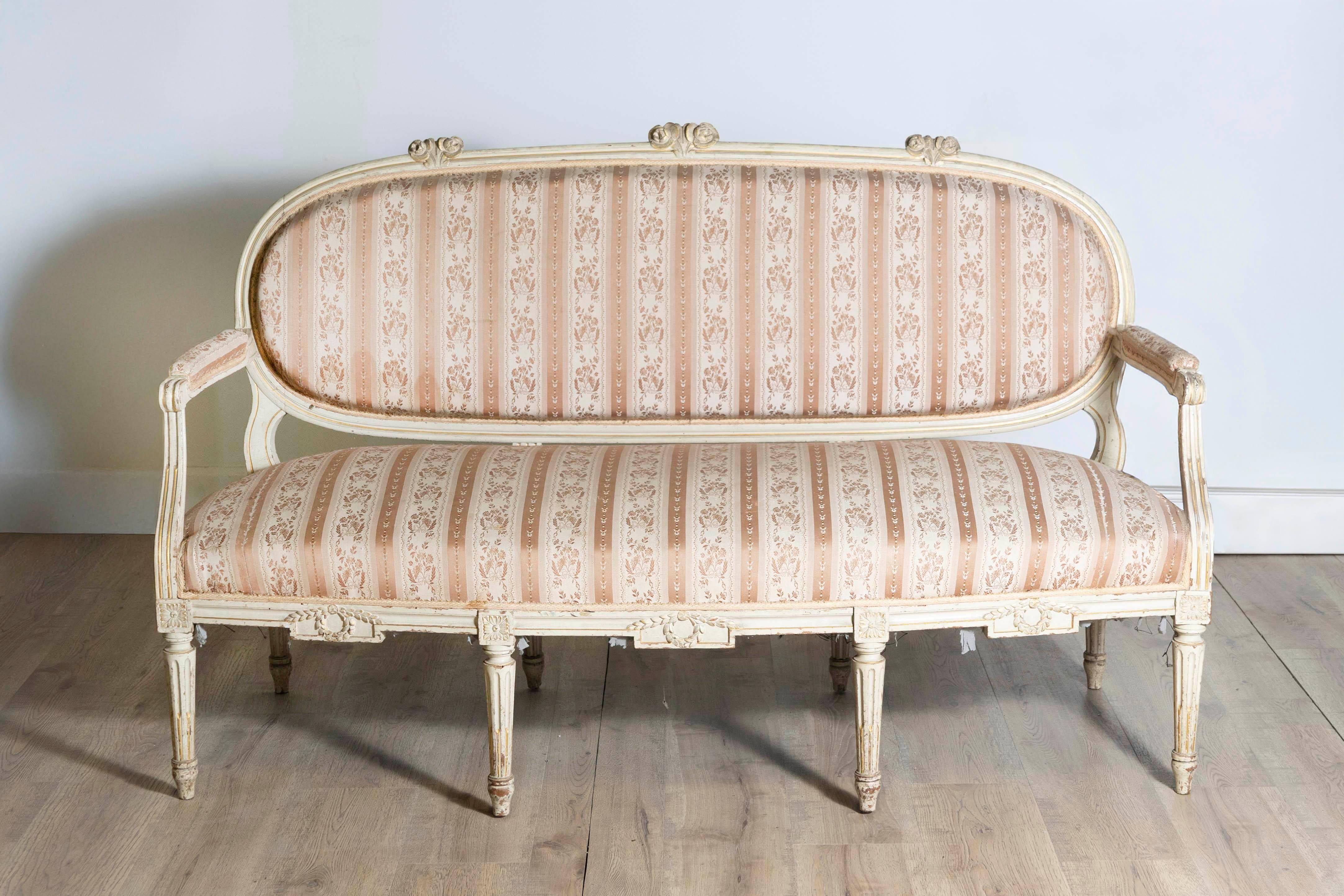 Canapé français d'époque Louis XVI, vers 1790, avec finition peinte en beige antique, dossier ovale, feuillage sculpté et pieds cannelés. Laissez-vous séduire par le romantisme d'une époque révolue avec cet exquis canapé français d'époque Louis XVI,
