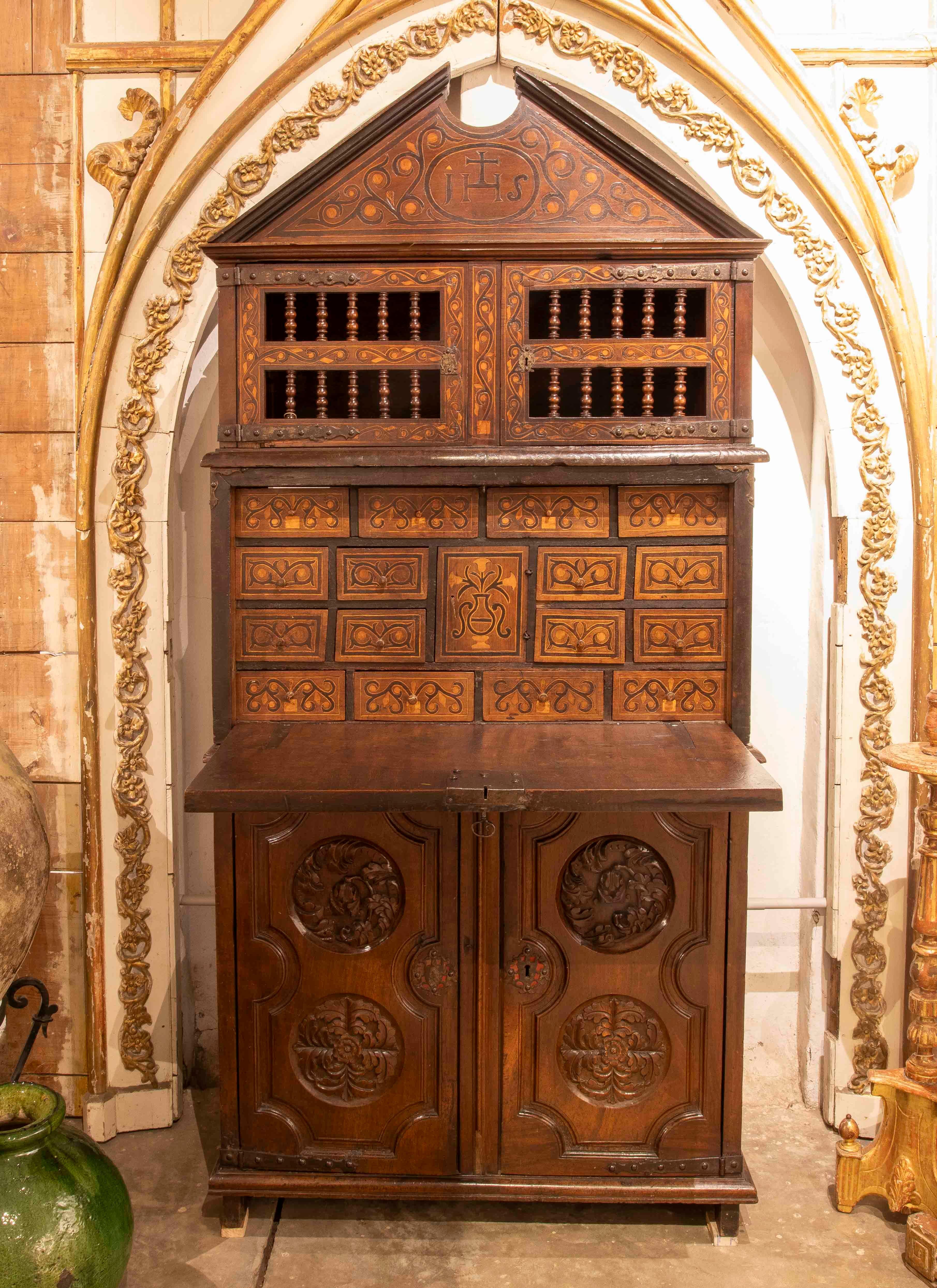 An impressive Asturian chest of drawers of Spanish origin, made of solid wood with lavish carved decoration and religious elements. The upper part features an architectural structure with a triangular pediment and the engraved IHS monogram, a