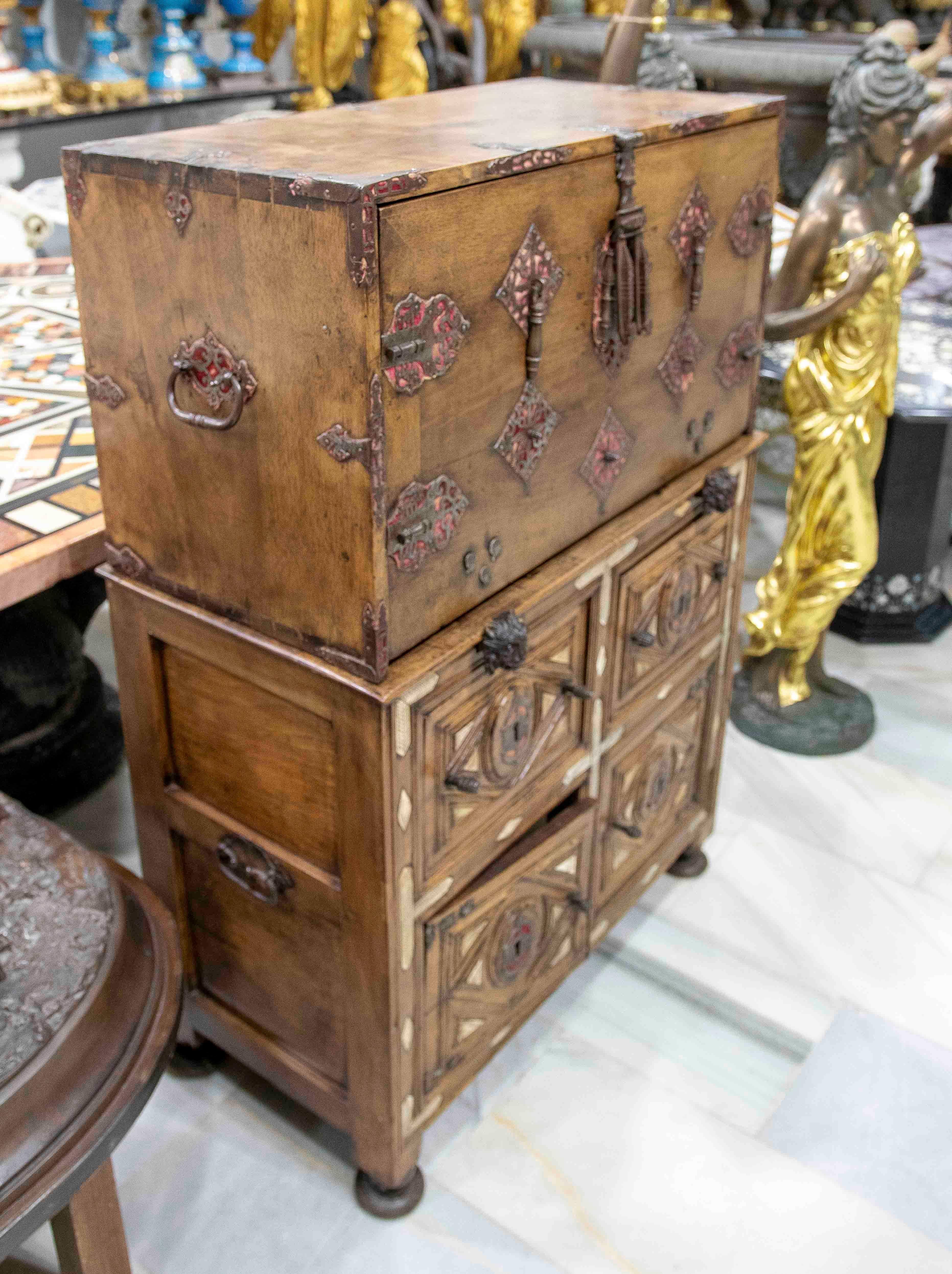 Impresionante conjunto formado por un bargueño de madera sobre un arcón a juego. El arcón presenta herrajes ornamentales de hierro forjado decorados en rojo, que contrastan con la calidez de la madera. Sus bisagras, cerradura y clavos decorativos