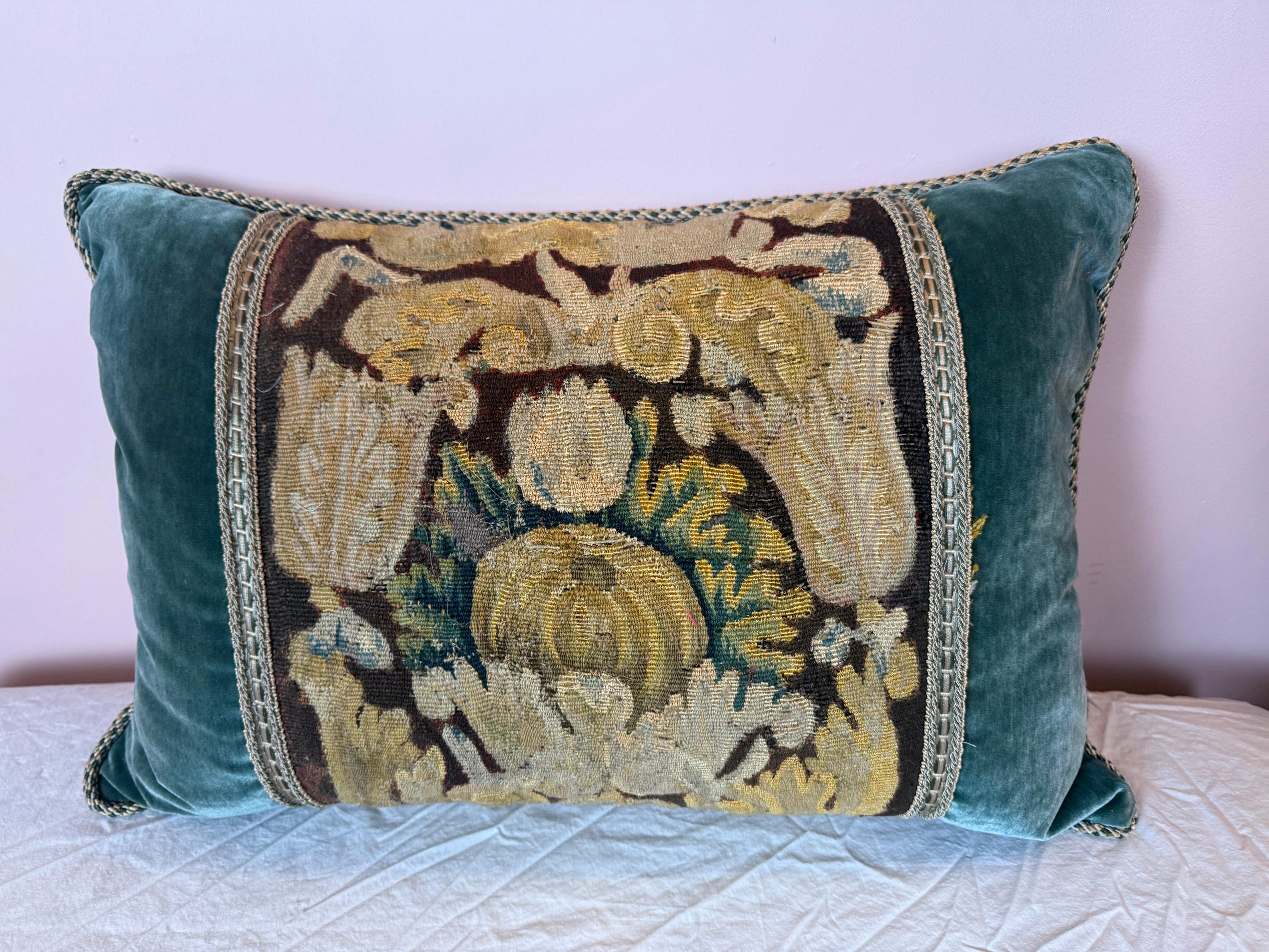 This elegant decorative pillow features a richly detailed 17th century tapestry fragment as its centerpiece, showcasing intricate florals and foliage in muted gold, green, and ivory tones against a dark ground. The antique textile is framed by