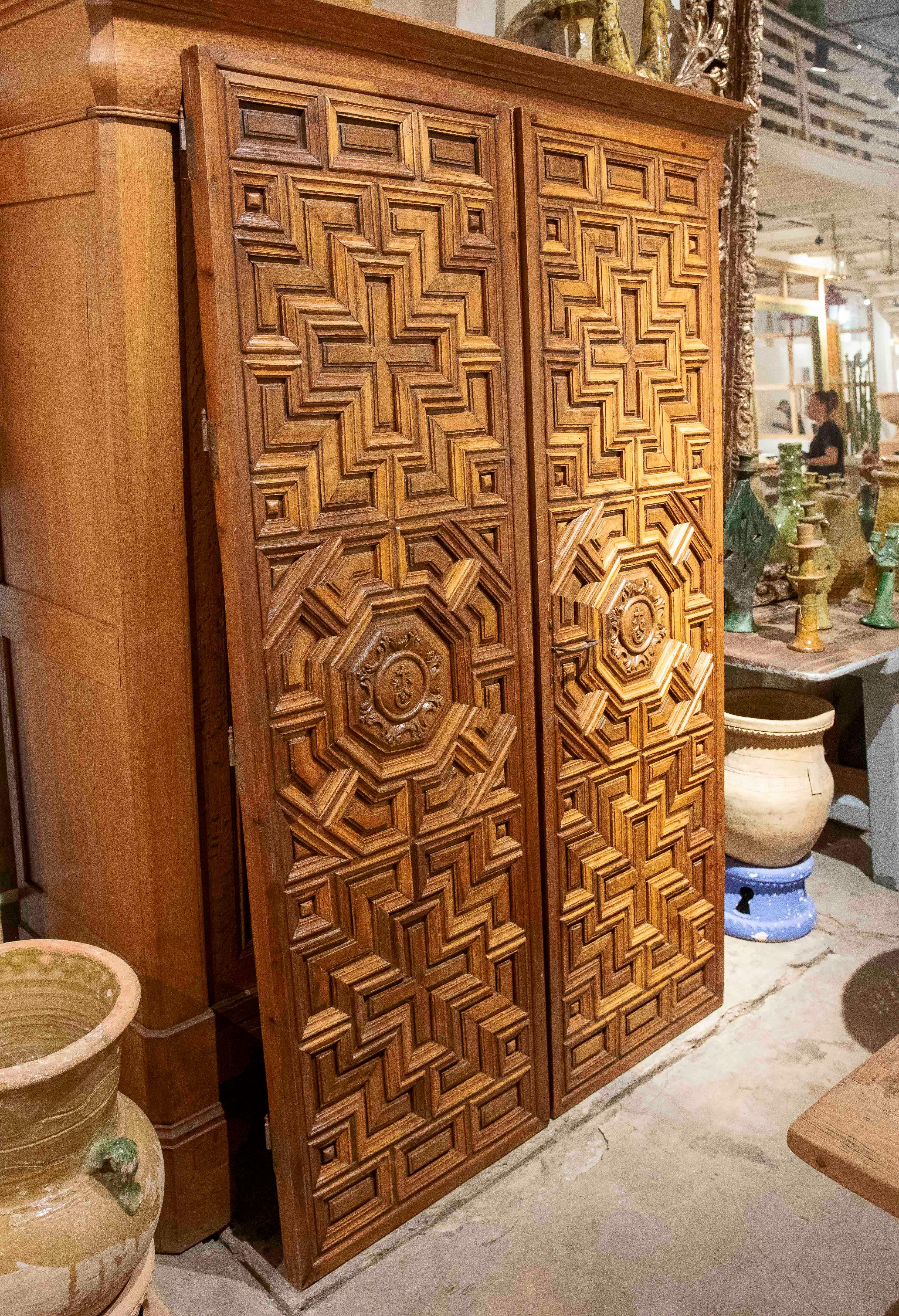 Double-leaf door crafted from solid wood, with rich relief ornamentation inspired by the Mudejar style. The design is composed of meticulously carved geometric patterns in a staggered perspective, creating a three-dimensional and symmetrical visual