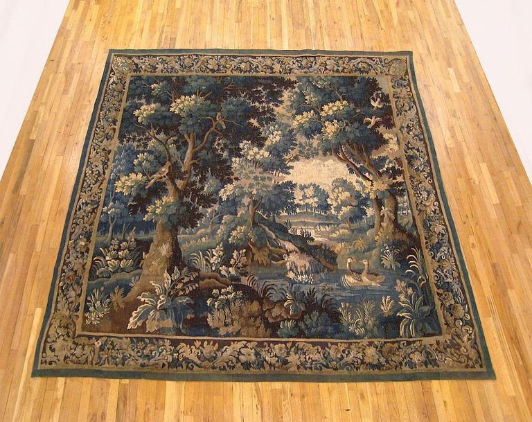 An Aubusson verdure landscape tapestry from the 17th century, depicting two ducks in a stream in the foreground amongst bird-filled trees in the woods, with rolling hills in the distance. Enclosed by a foliate border with acanthus leaves and
