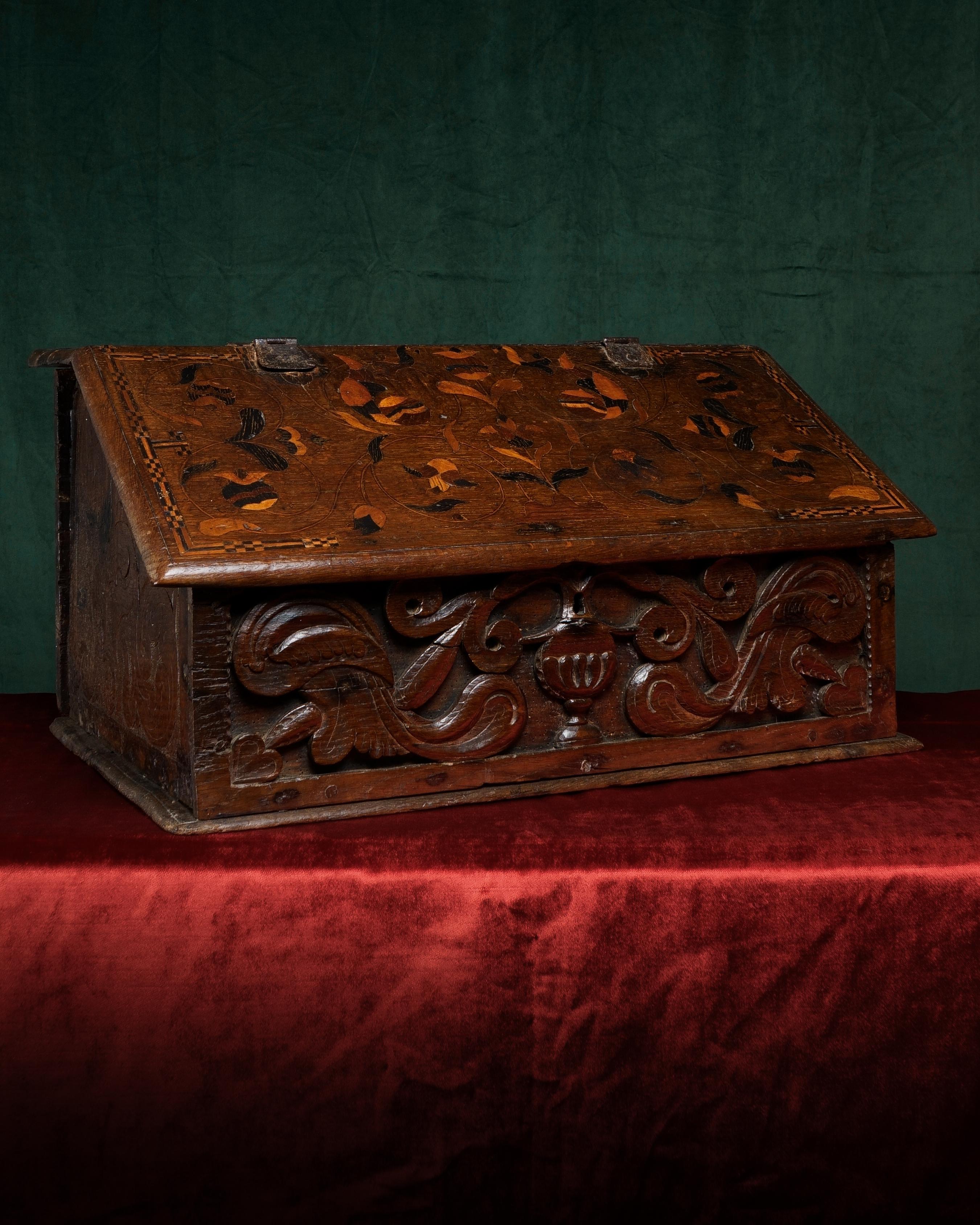 The top board bearing marquetry-inlaid initials ‘MA’, the slope with further marquetry-inlaid flowers and meandering foliage, enclosing an interior with a shelf, the end boards boldly carved with foliage, the front board carved with a flower-filled