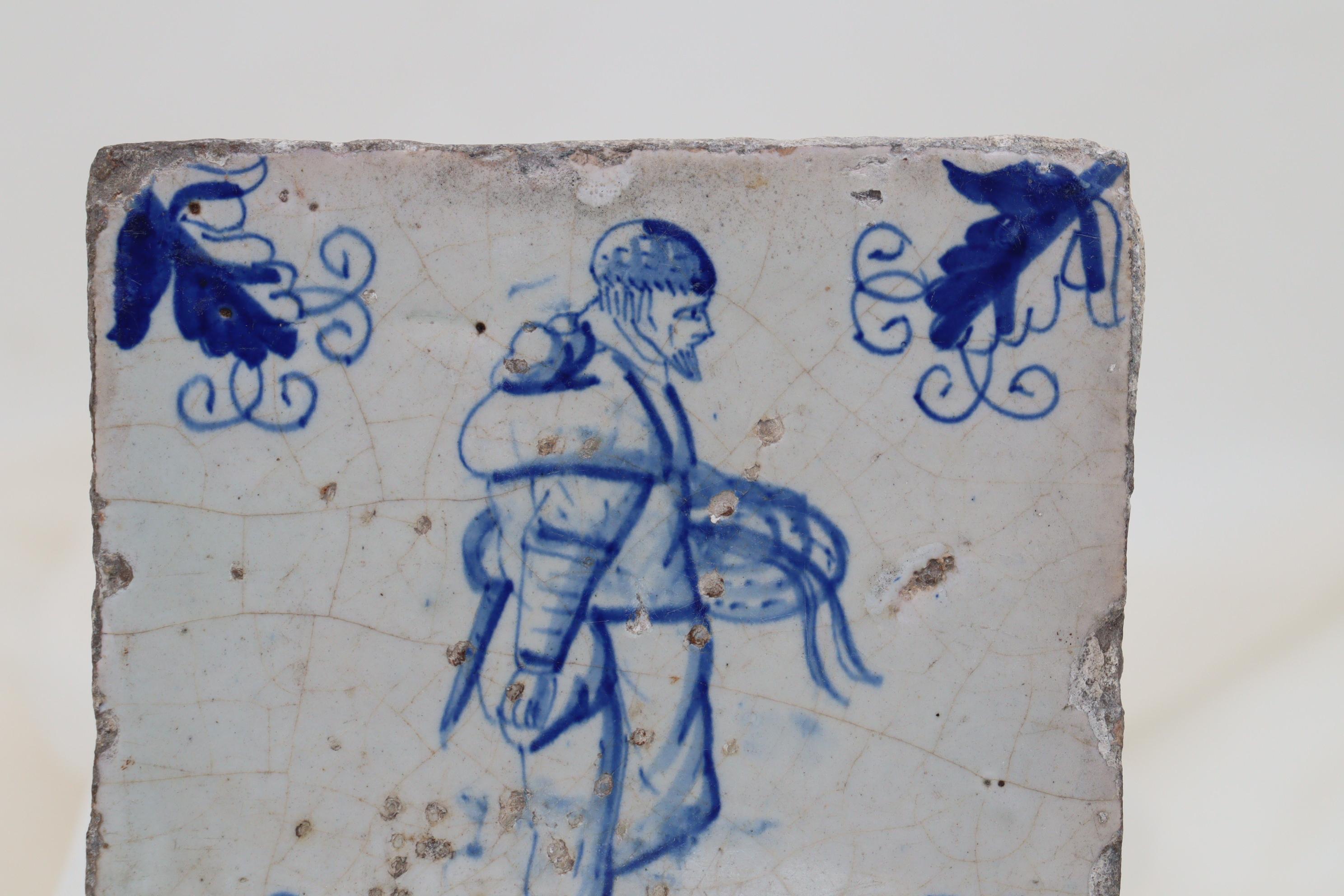 Diese holländische Delft-Kachel aus dem 17. Jahrhundert zeigt in der Mitte die handgemalte Figur eines Mannes, der einen blauen Korb trägt, und an den Ecken Motive von Kriechblättern. Die Figur könnte einen Korb mit Aalen tragen, die in den
