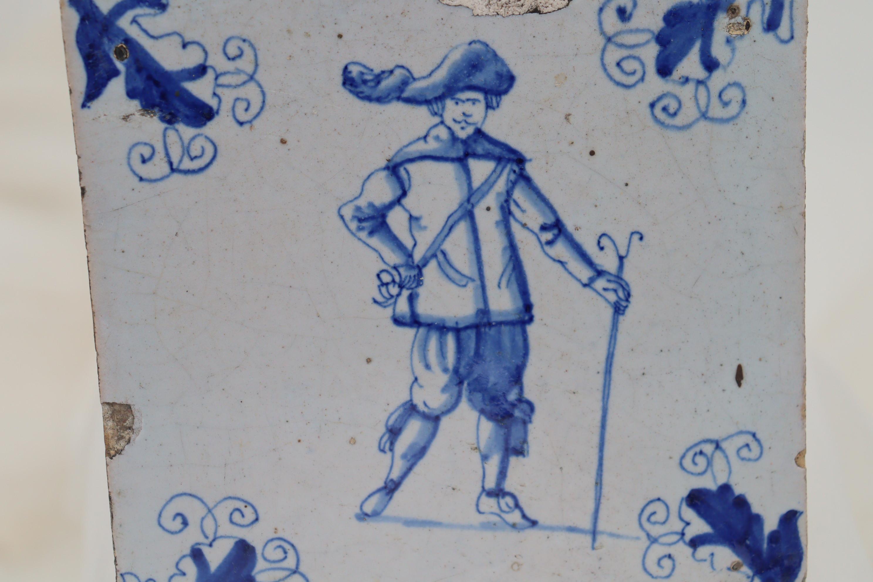 In the centre of this Dutch hand painted delft tile is a portrait of a well dressed man of the 17th Century carrying a staff or stick. To the corners are creeper leaf motifs, and interestingly, on the rear of the tile, is the drawn outline of a
