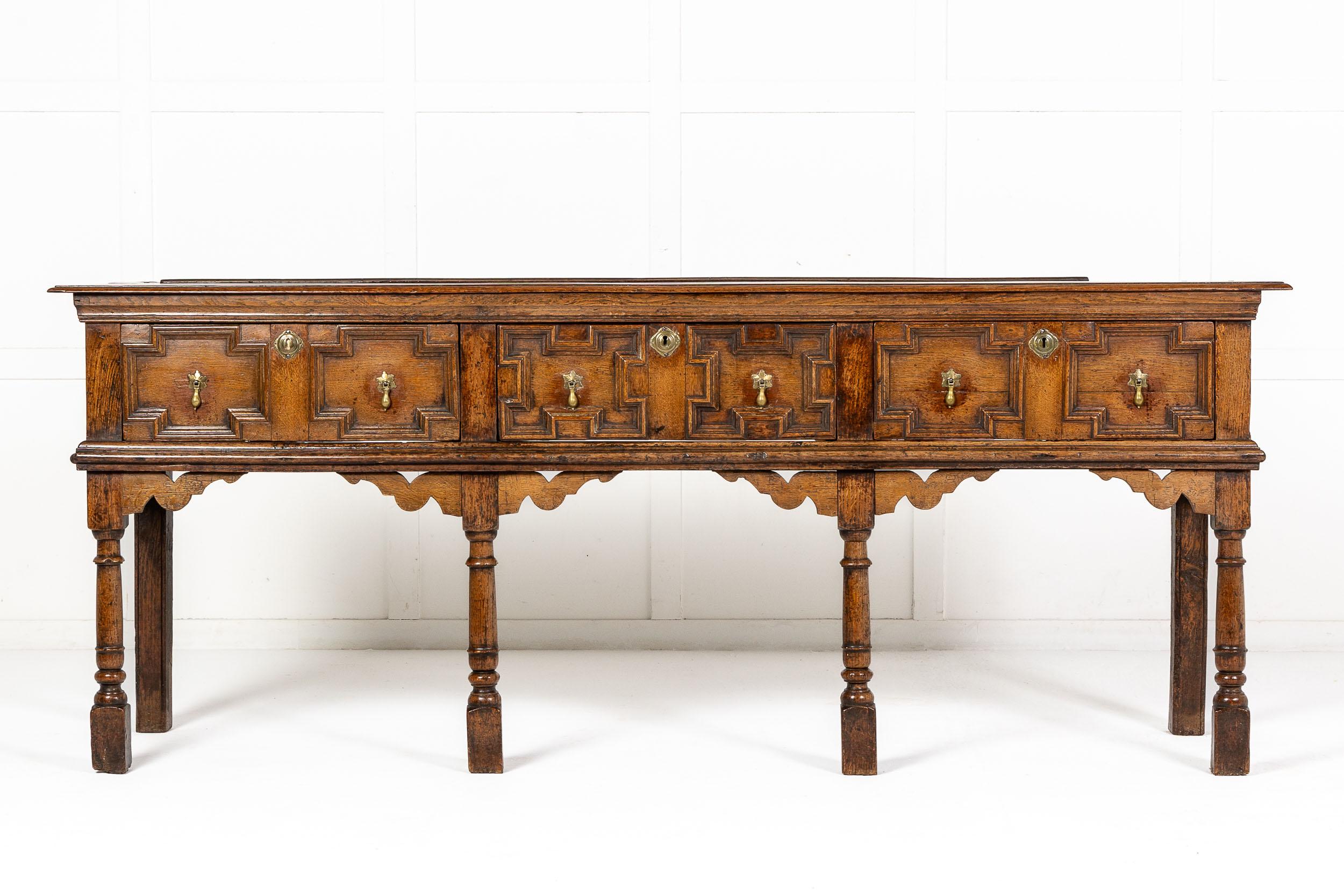 17th century English oak dresser base with a moulded edge, plank top and three deep drawers below with excellent moulded panels as geometric decoration, brass pull handles and escutcheons. Supported by four sturdy, turned front legs united by