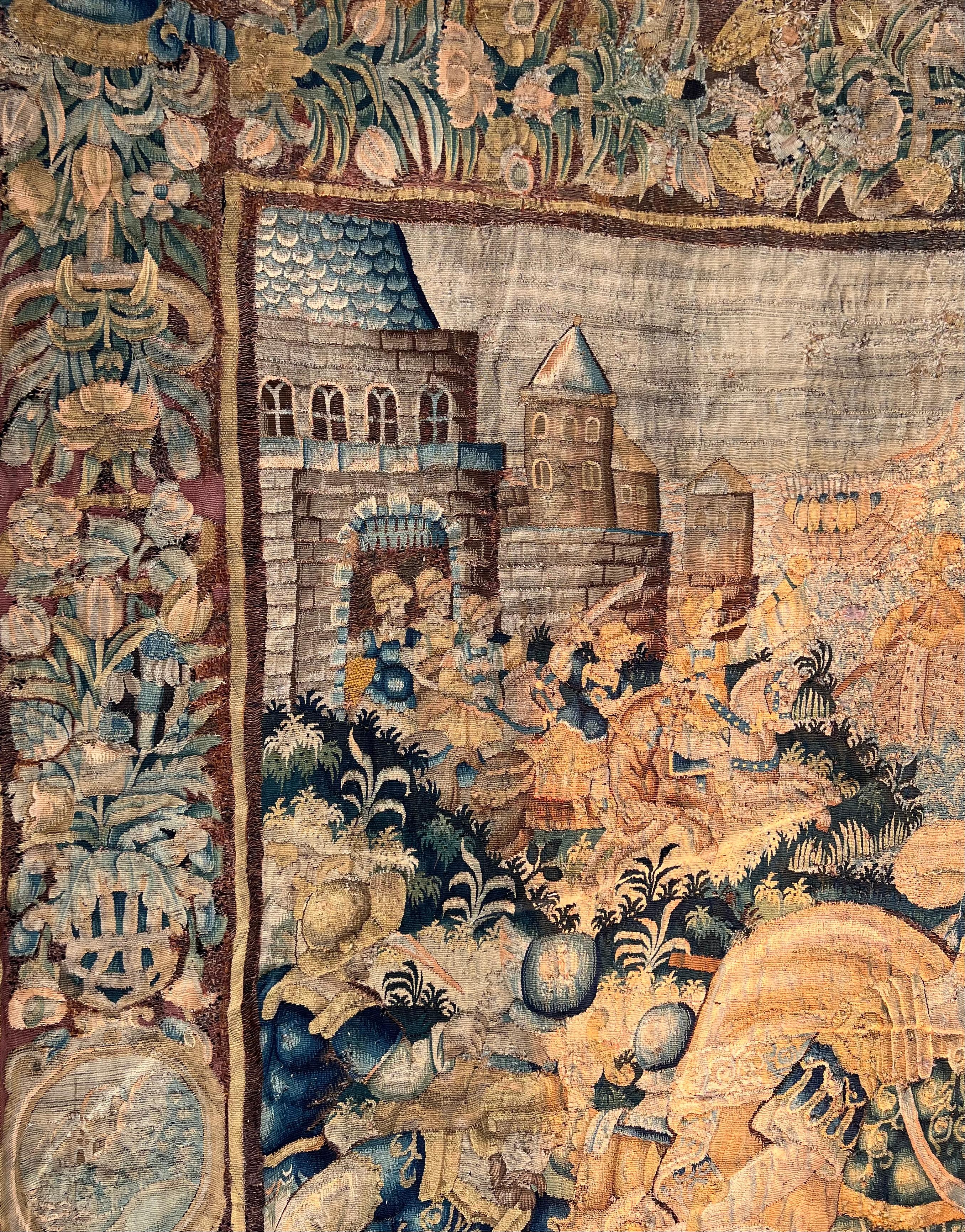 A large palatial Flemish baroque historical tapestry depicting a battle scene, with soldiers to the foreground on land, the opposing army arriving by sea, with a city under siege to the distance, within a border of floral and foliate ornament.