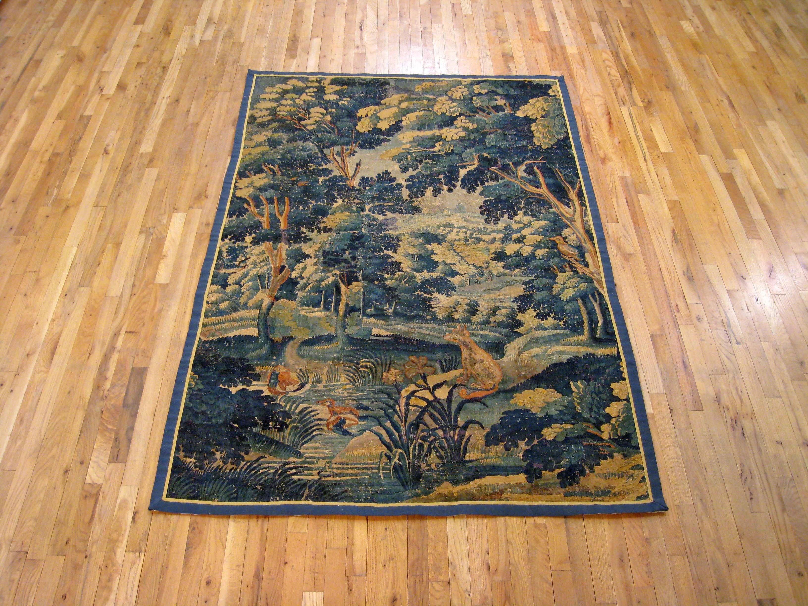 A Flemish verdure landscape tapestry from the 17th century, featuring a fox reposing by a pond in a verdant forest setting, with small bushes in the foreground, large trees in the middle distance, and further greenery in the background. Enclosed