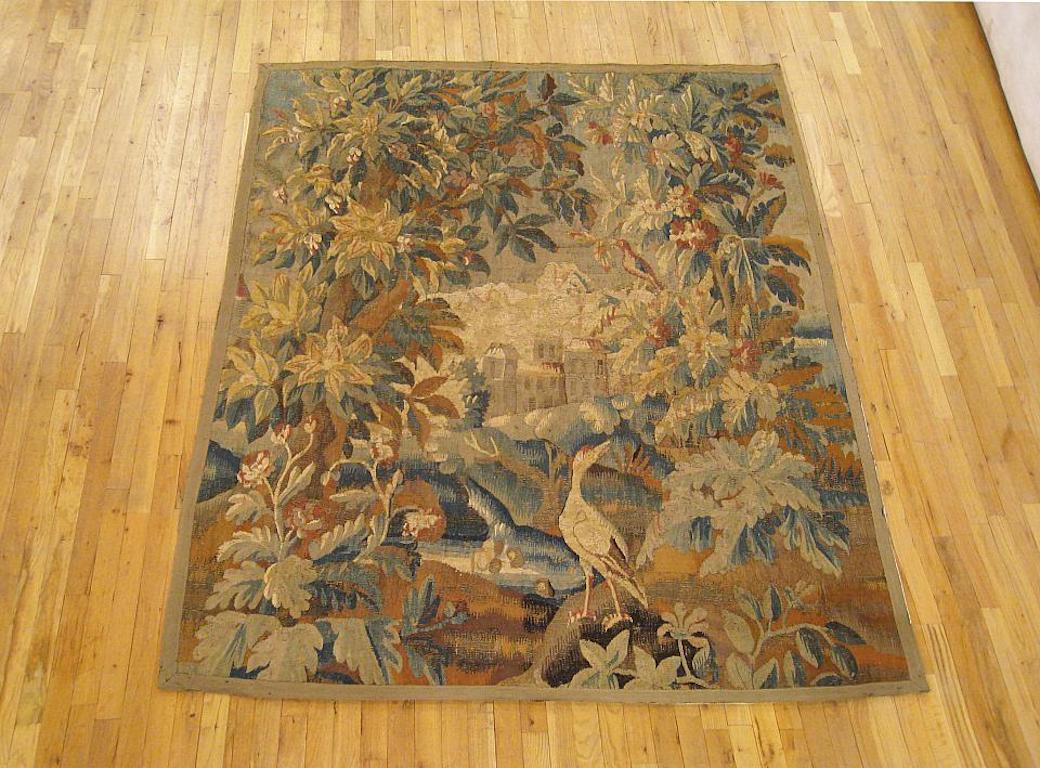 A Flemish verdure landscape tapestry from the 17th century, envisioning an idyllic scene with a heron on a grassy bank between various flora and trees in the foreground, with a pond in the middle distance and a stately manor amidst the rolling hills