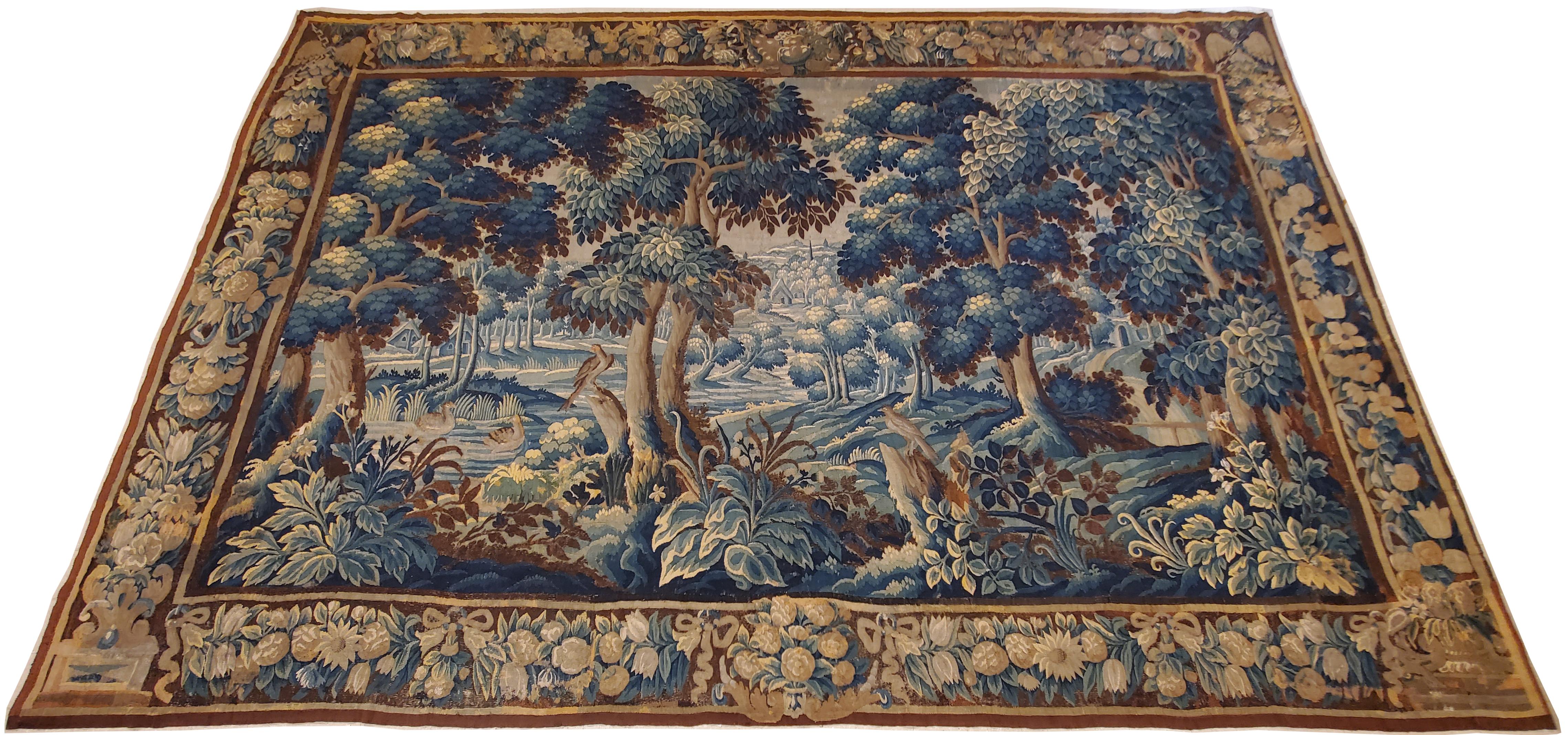 Large Flemish verdure tapestry from the 17th century, featuring a tranquil scene in a woodland setting, with stately trees at right and left in the foreground, with an exotic bird resting in the grass beneath their shadow, and further trees in some
