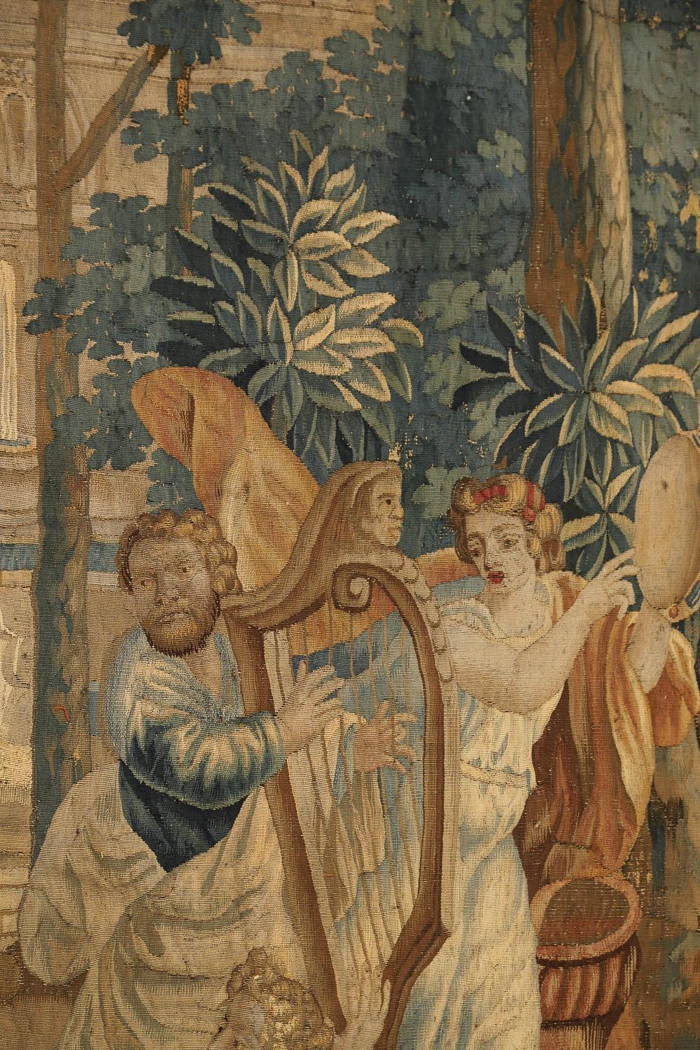 Flemish Verdure Tapestry with Classical Figures as Musicians, 17th Century.
Rectangular in shape, the colorful piece depicts an outdoor scene near a fountain with a man playing a harp while one woman plays a tambourine, and another woman lounges