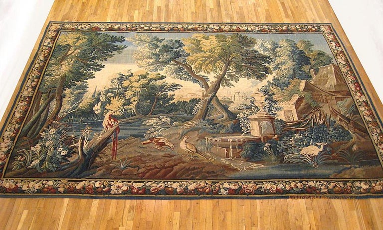 A French Aubusson verdure landscape tapestry from the 17th century, featuring a paradisiacal vision of a remote cottage with a beautiful waterfall placed within a lush lakeside setting, replete with various trees, flowers, and exotic birds, and with
