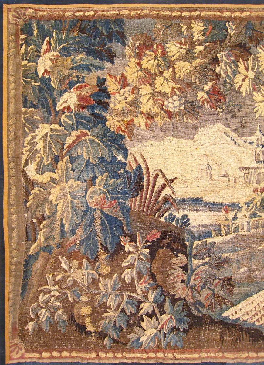 An antique 17th Century French Verdure Landscape Tapestry, envisioning a scene in which a dog chases an exotic bird at left, and a villagescape is visible in the middle distance.  There is a large tree branch extending at the top end of the central