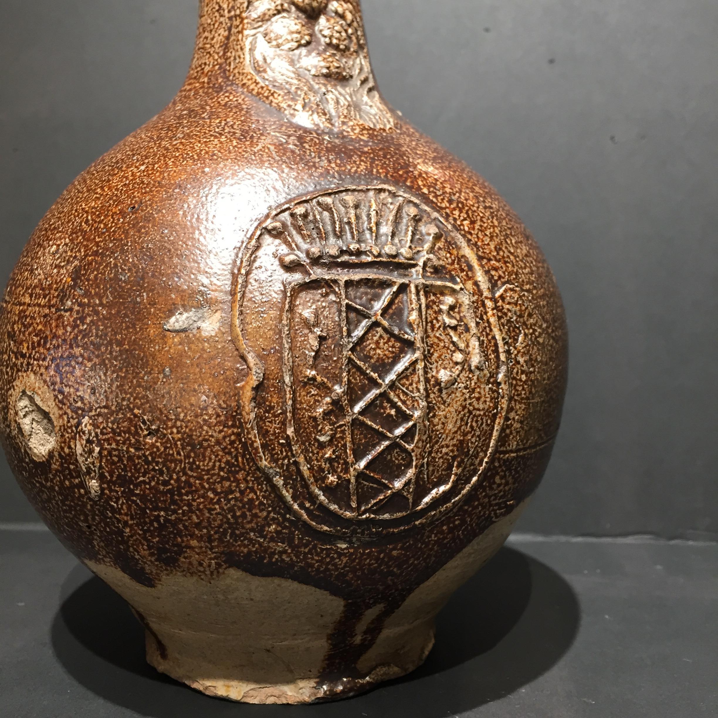 Germany
Frechen
Circa 1620 – 1640

Bellarmine or Bartmann jug with a coat of arms.

A salt-glazed and iron-engobed bellarmine with a narrow neck, spherical body, and flat base. A stylized face with a closed mouth and a long round beard is applied to