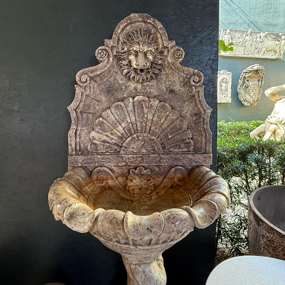 Fuente mural de mármol con pedestal de delfín. El cuerpo en forma de campana de la jofaina se apoya en el pedestal grotesco delfín, que está colocado sobre un pie cuadrado elevado. El fondo de la fuente está decorado con volutas y una cabeza de león