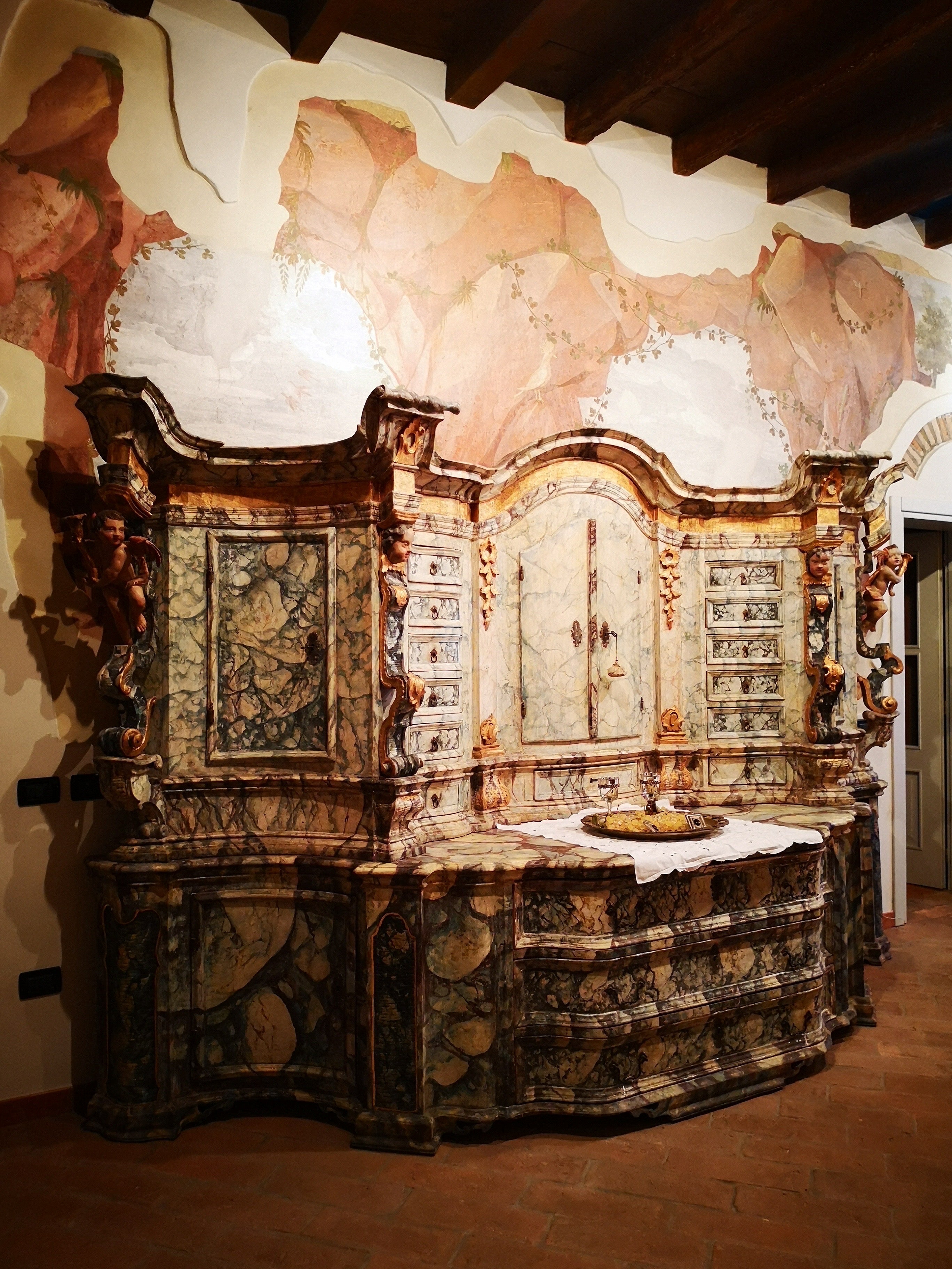 Este monumental mueble, de procedencia véneto-alto véneto, está fabricado íntegramente en madera de abeto lacada. Pleno siglo XVII (hacia 1650) se presenta como un imponente aparador de doble cuerpo, enteramente lacado en falso mármol con diversos