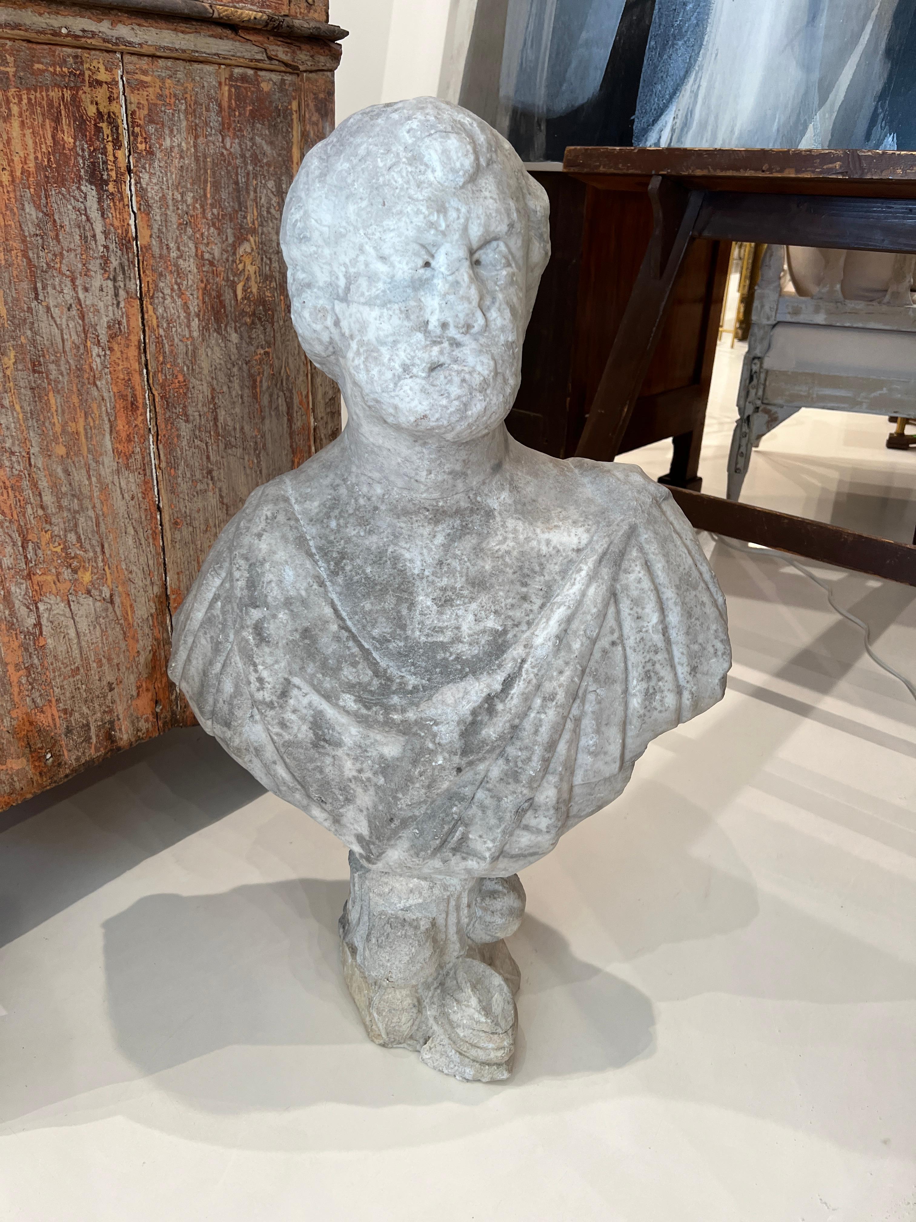 Beautiful, albeit heavily distressed stone bust depicting an authoritarian man on a pedestal.  Softly colored grey stone with many details intact including draping on tunic, eyes, beard and floral motifs at base.  
Mounted on a pedestal at eye level