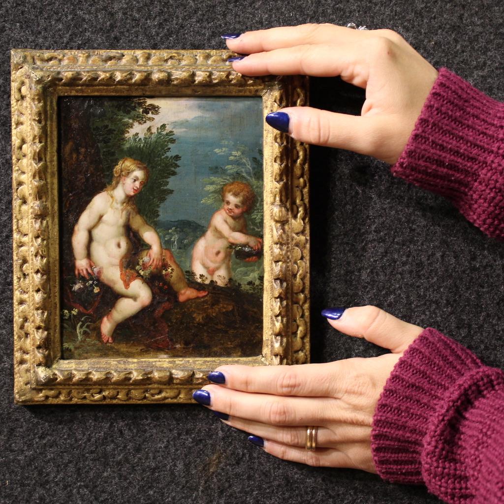 Una refinada pintura flamenca del siglo XVII. Este óleo sobre cobre representa una espléndida escena mitológica, un maravilloso paisaje con Venus y un cupido. La joven diosa está sentada al pie de un árbol, tejiendo y recibiendo flores de un