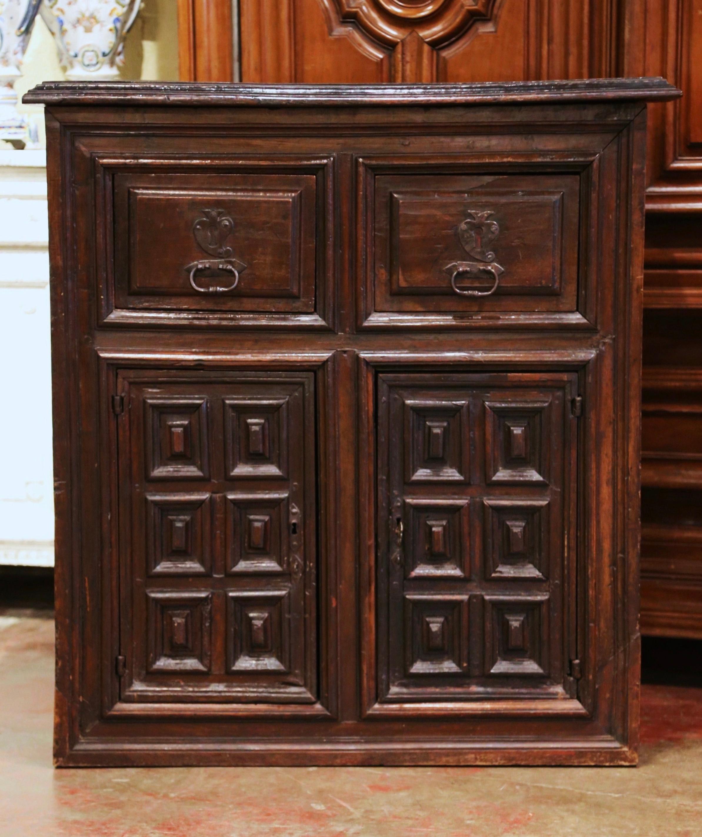 Hand carved in northern Spain circa 1700, the fruit wood cabinet features two drawers across the front dressed with the original forged pulls and escutcheons over two intricate hinged doors decorated with geometric square raised panels on both