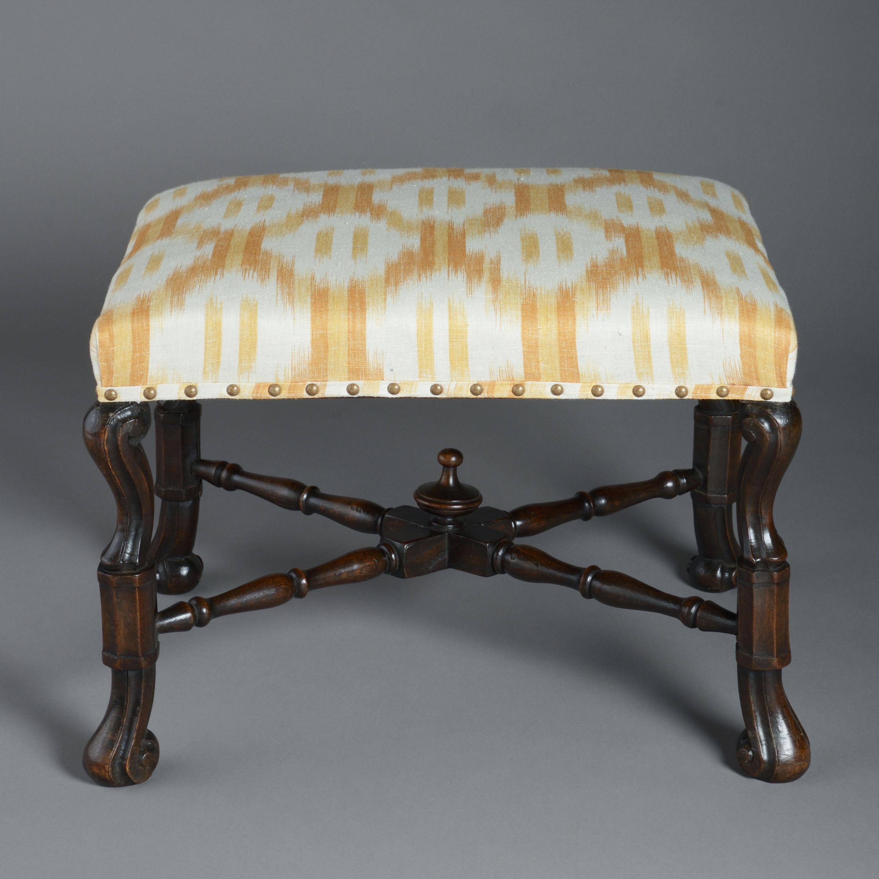 A late 19th century walnut stool in the Charles II manner, the upholstered seat upon carved braganza legs with a conjoining X-form stretcher, surmounted with a turned finial.