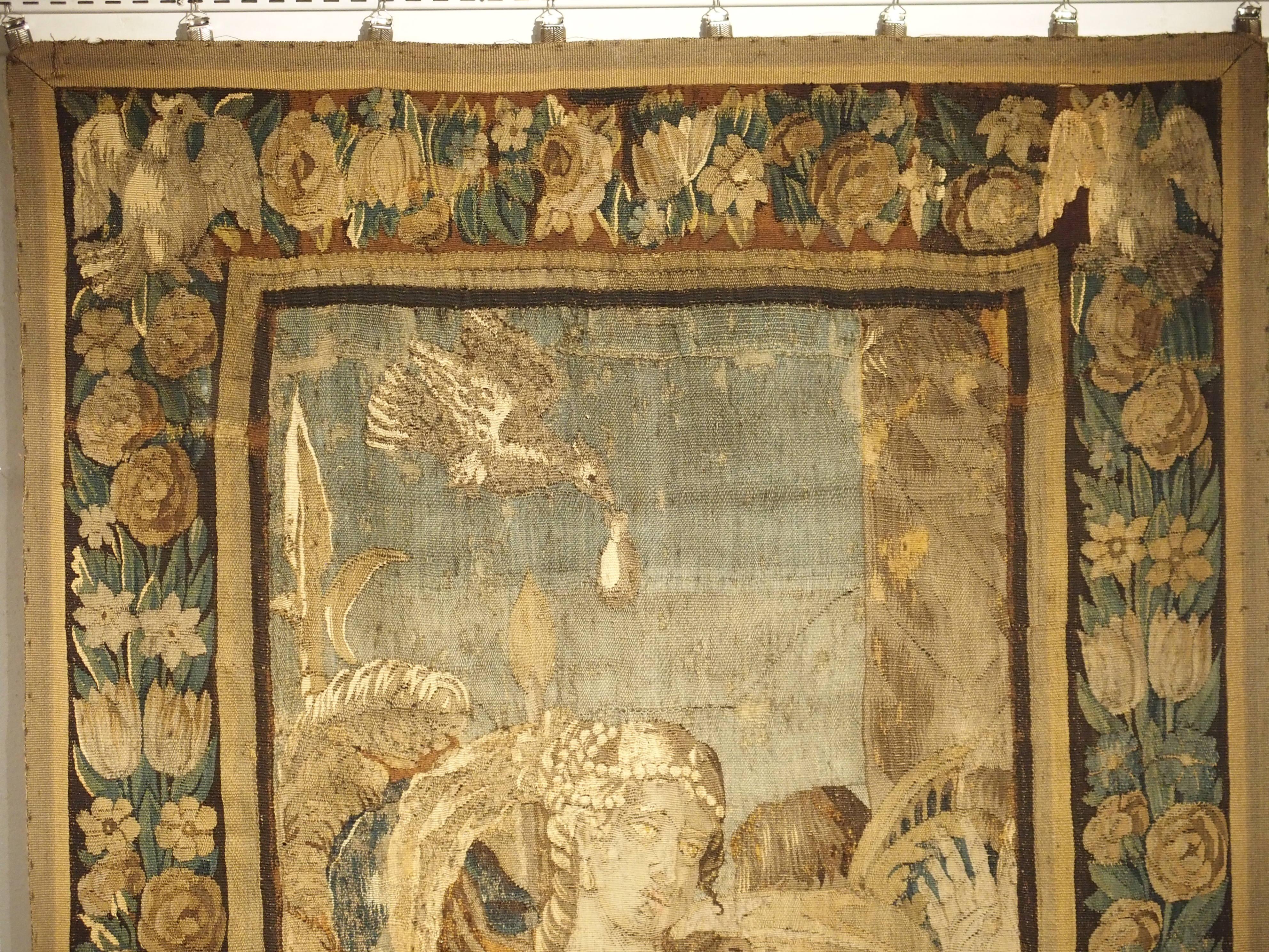 This incredible tapestry fragment depicts a Noble woman with a warrior standing to the back left, carrying a halberd. She has pearls in her hair and on her wrist, and is wearing a robe trimmed in ermine and adorned with Fleur de Lys throughout. A