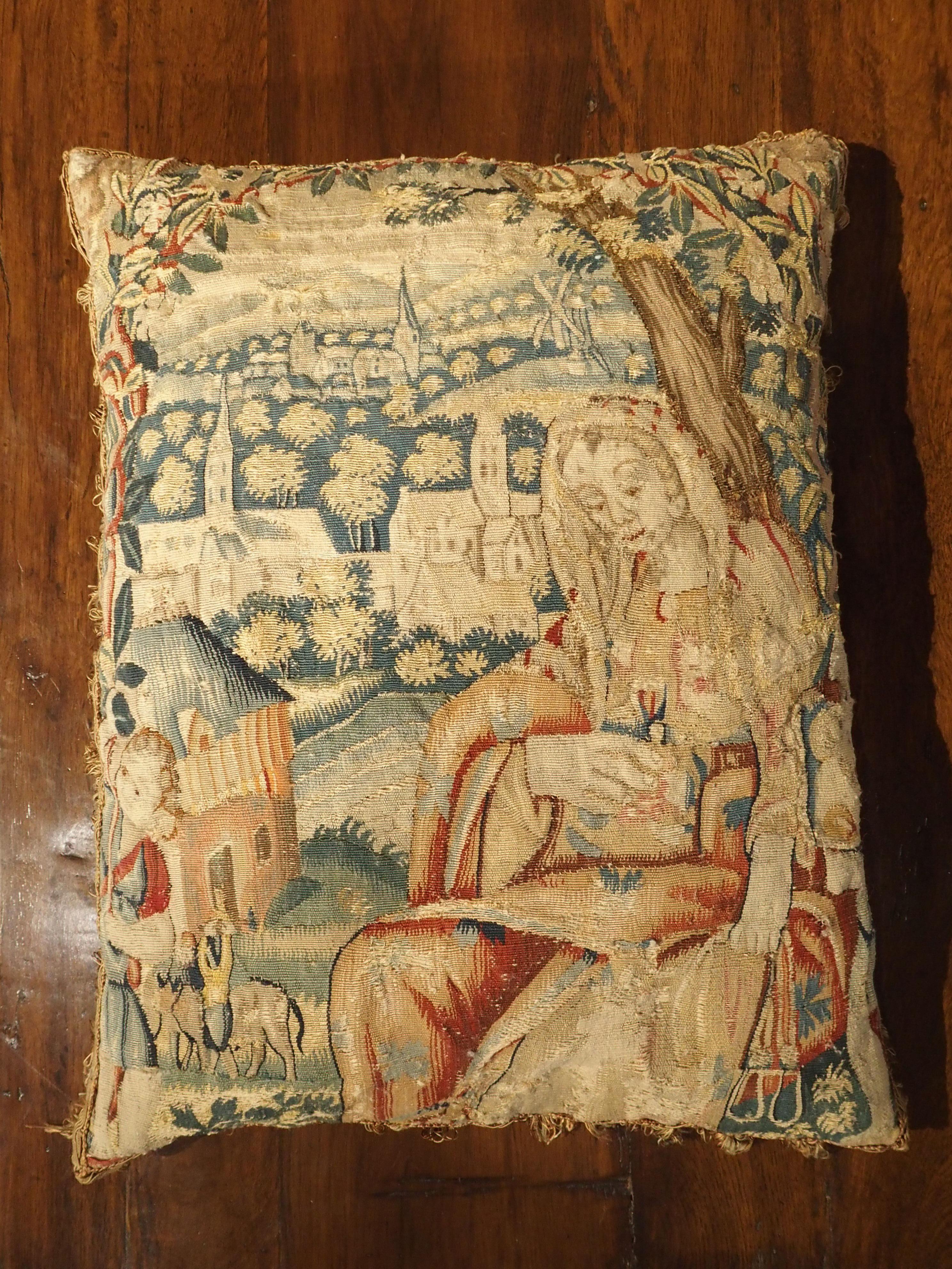 From France, this beautiful tapestry pillow dates to the 1600s. The scene depicts a woman in the foreground, sitting under a tree with stirrups in her hand. On the left side of the pillow, as the scene fades into the background, we see smaller