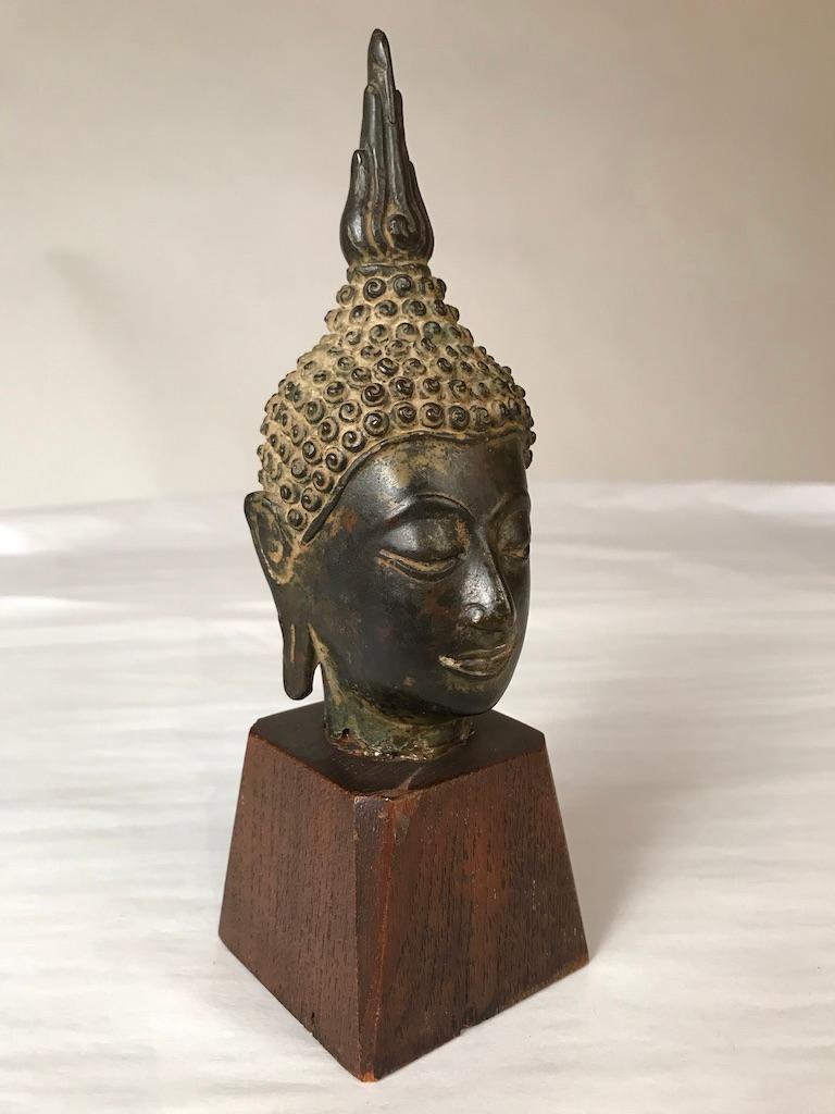 A Thai, Ayutthaya style, bronze head of Buddha Shakyamuni, late 16th or early 17th century. With a serene expression on his face, downcast eyes, arched eyebrows, smiling lips, with curled headdress and ushnisha topped by a flame. 
Measures: 7.75