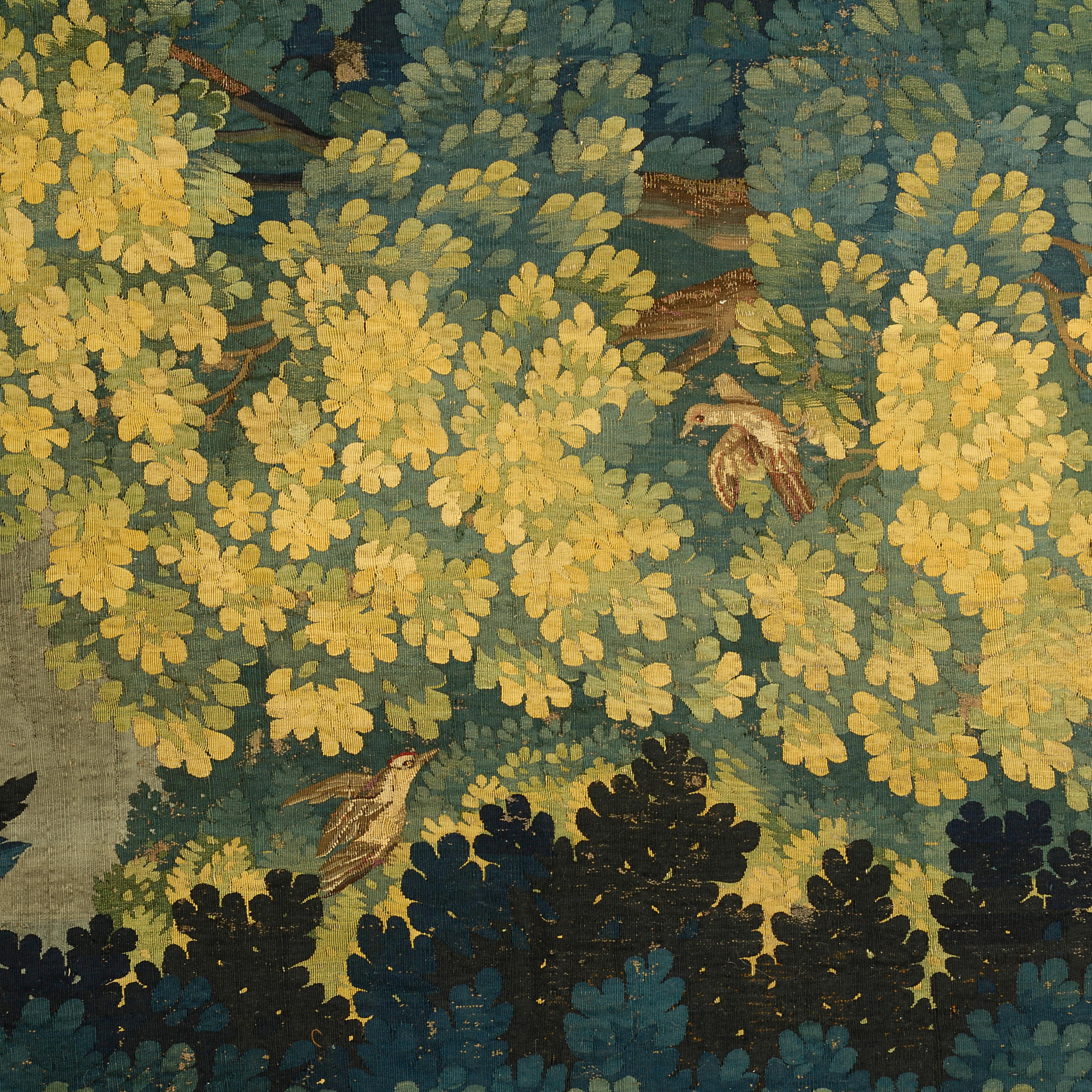 Tapisserie en soie et laine finement tissée, représentant un paysage boisé, avec un oiseau posé sur un tronc d'arbre et des plumes d'autruche dans chaque coin.