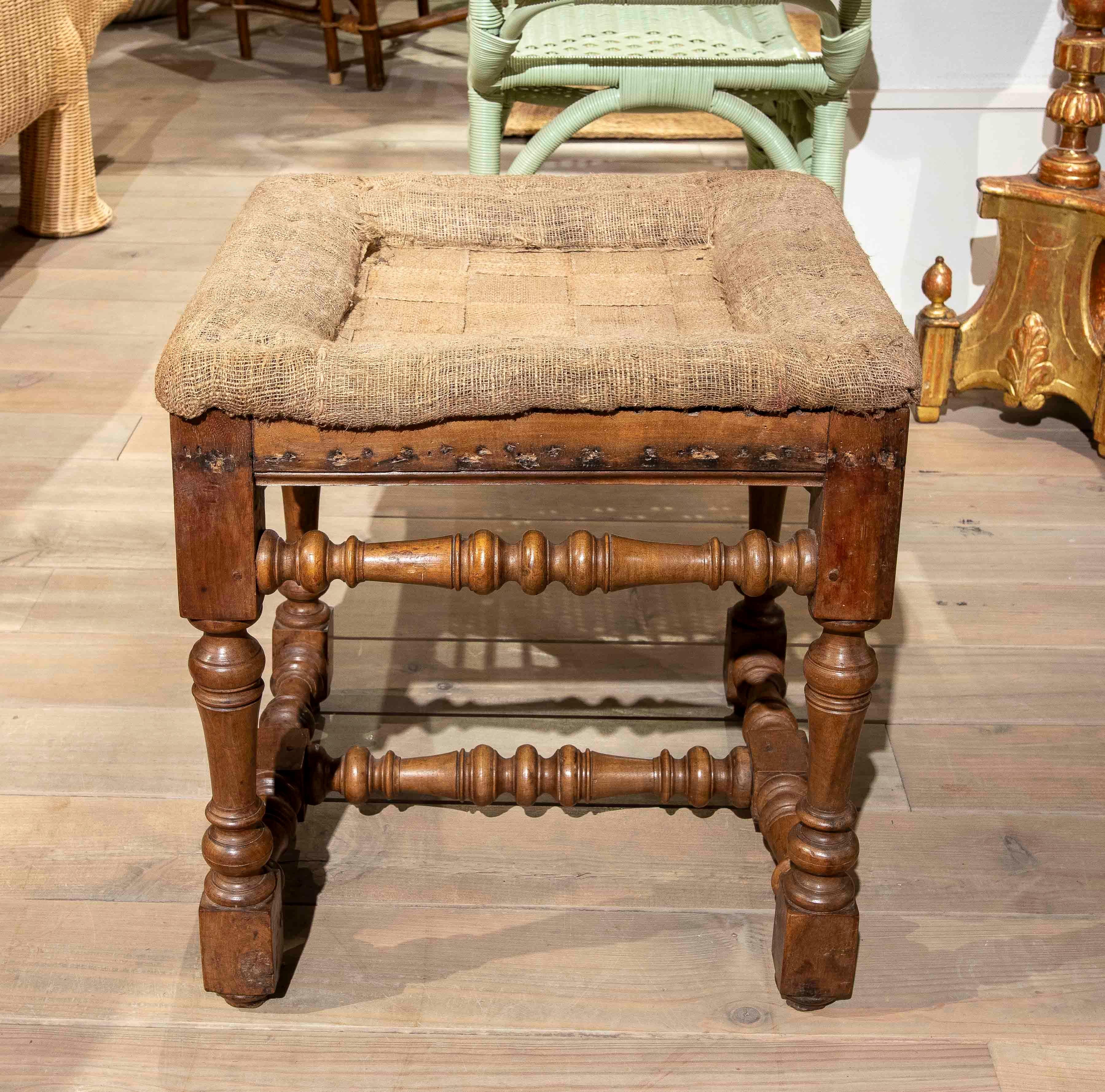 Charmanter Hocker mit geschnitztem Massivholzrahmen, gedrechselten Beinen und dekorativen Querstreben, die ihm Charakter und Stabilität verleihen. Die Sitzfläche besteht aus natürlichem Stoff mit einem geflochtenen Muster, das einen rustikalen,