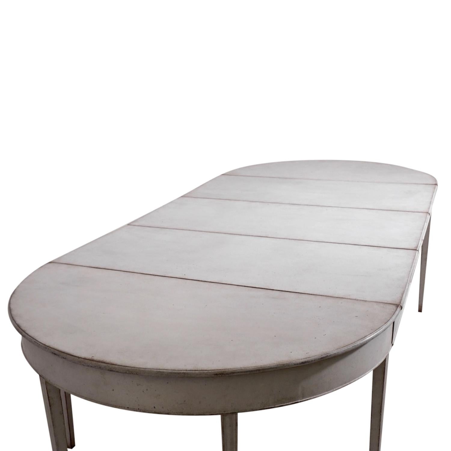 An antique Swedish Gustavian extendable dining table made of hand carved Pinewood with three leaves and apron. The light-grey, Scandinavian half round table is standing on nine straight square tapered fluted legs, detailed in the Neoclassical Greek