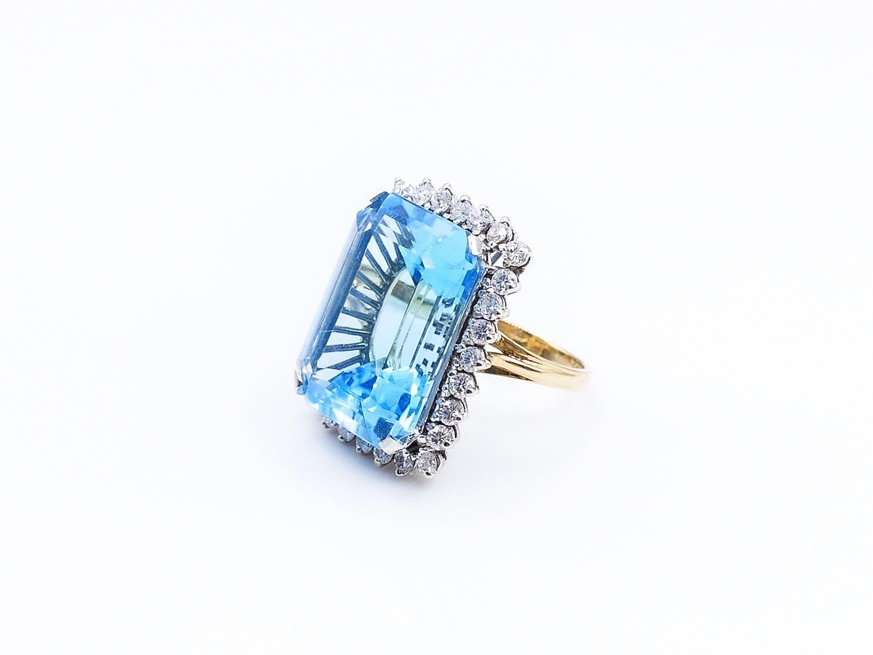 This bright and beautiful emerald cut London Blue Topaz, weighing in at 18 carats, is surrounded by 28 small round cut prong set diamonds totaling approximately 1.5 carats. The face of the ring is set in 18K white gold while the band is 18K yellow